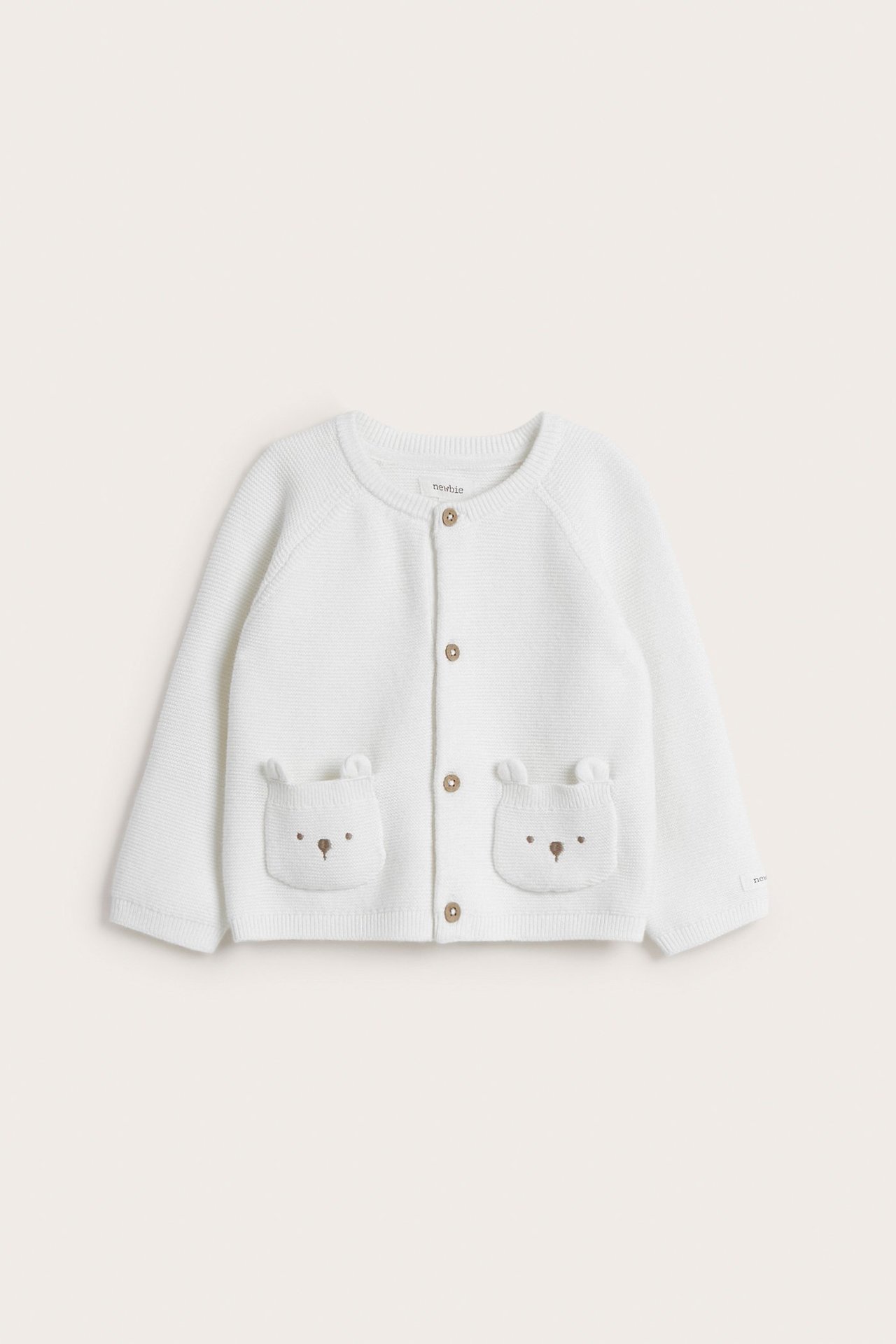 Frontvisning av en offwhite strikket babycardigan i bomull med knapper og to broderte bjørnelommer.