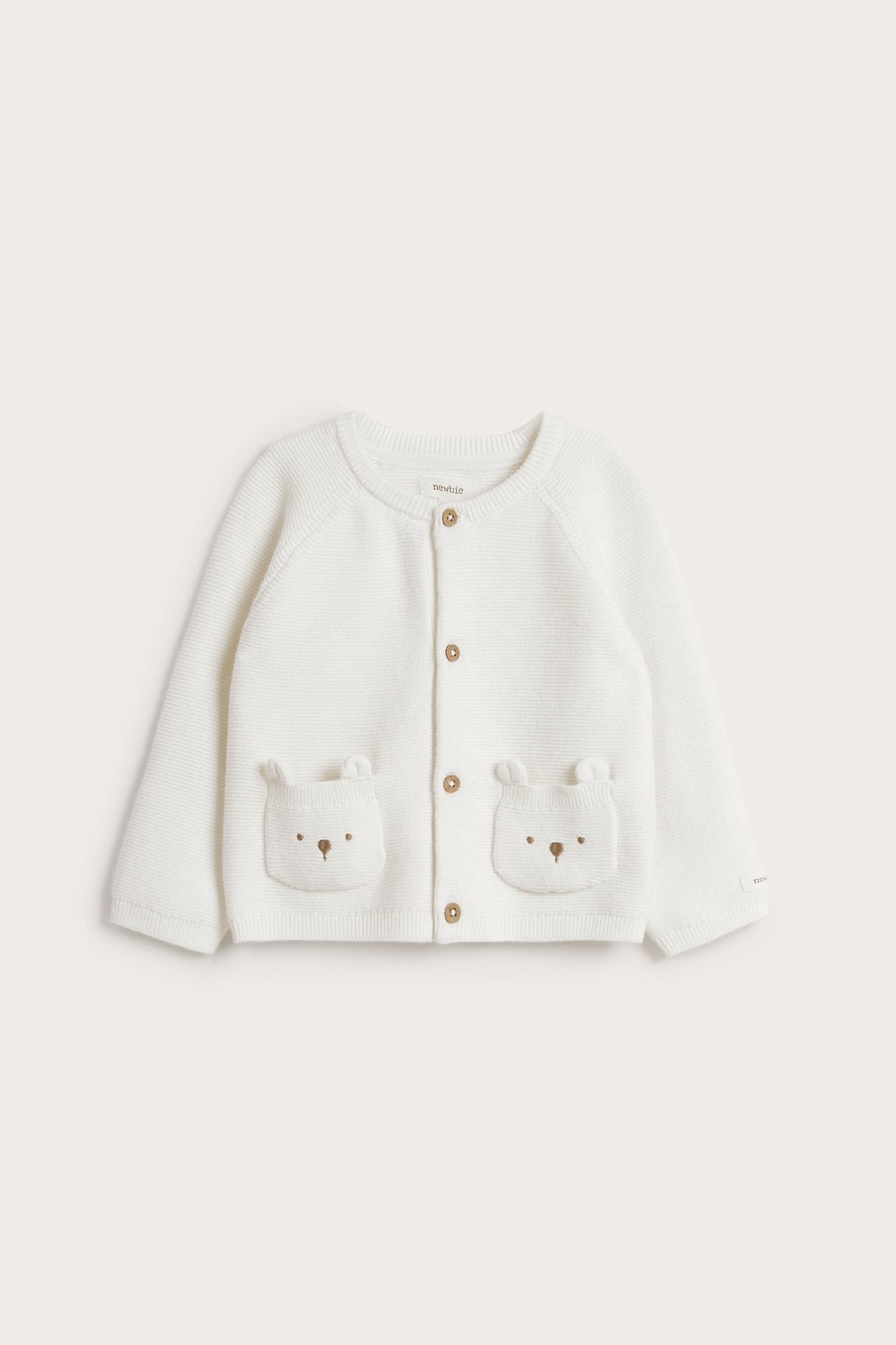 Frontvisning av en offwhite strikket babycardigan med bamseøre-lommer og brune knapper.