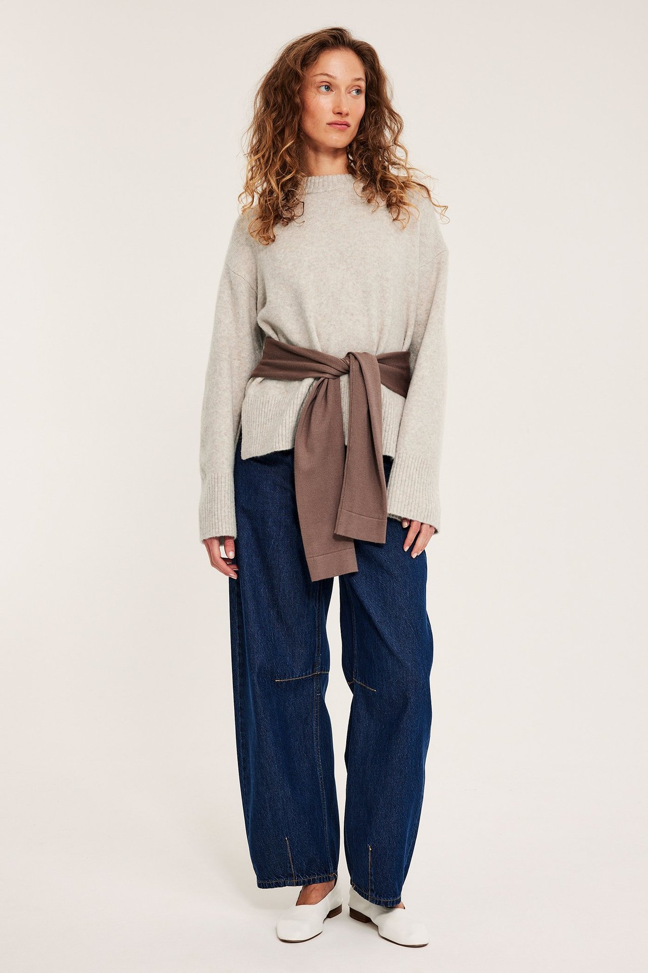 Nainen beigeessa pyöreäkaulaisessa neulepaidassa, siniset farkut ja valkoiset kengät.