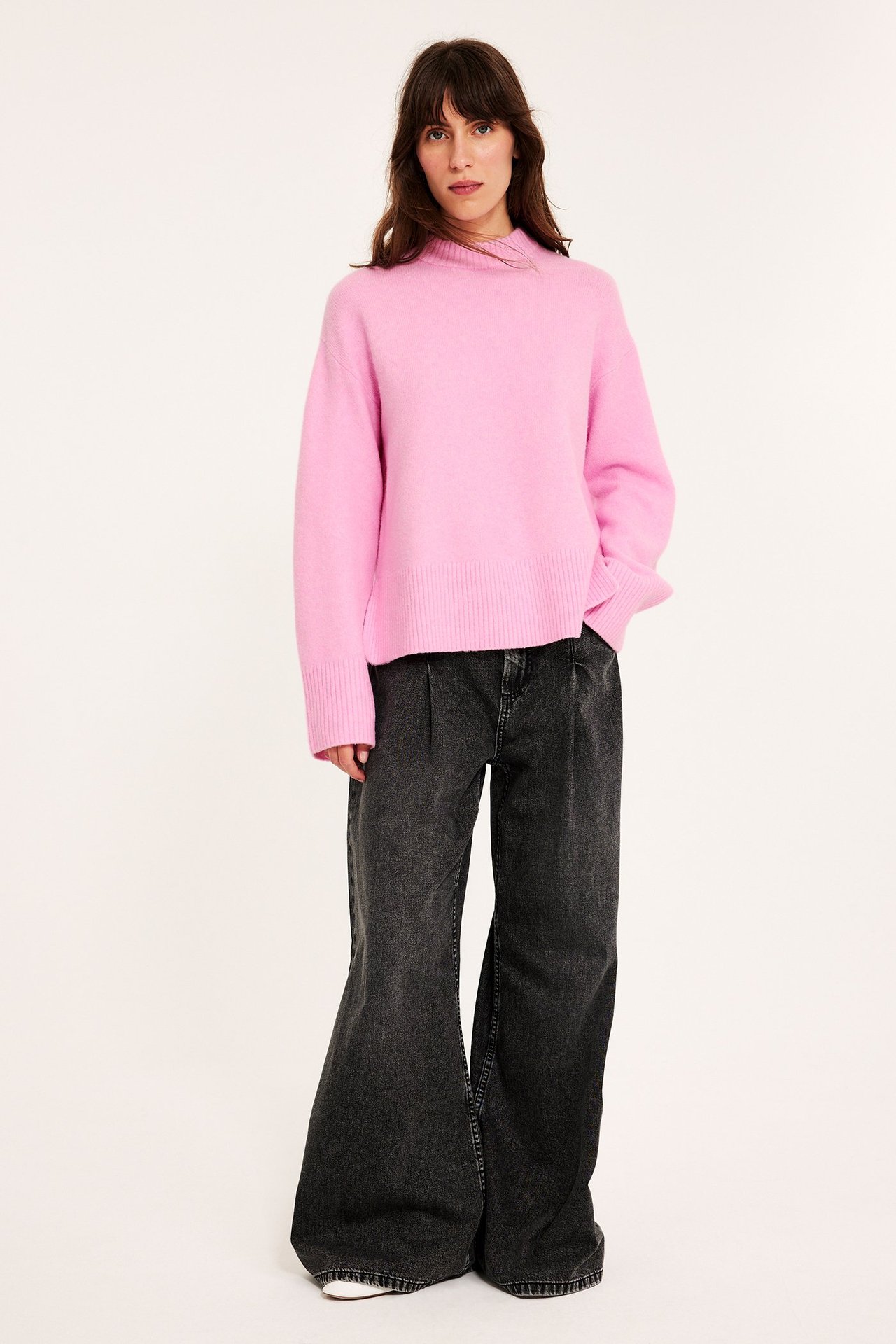 Edestä: nainen yllään vaaleanpunainen neulepusero ja tummanharmaat leveälahkeiset farkut.