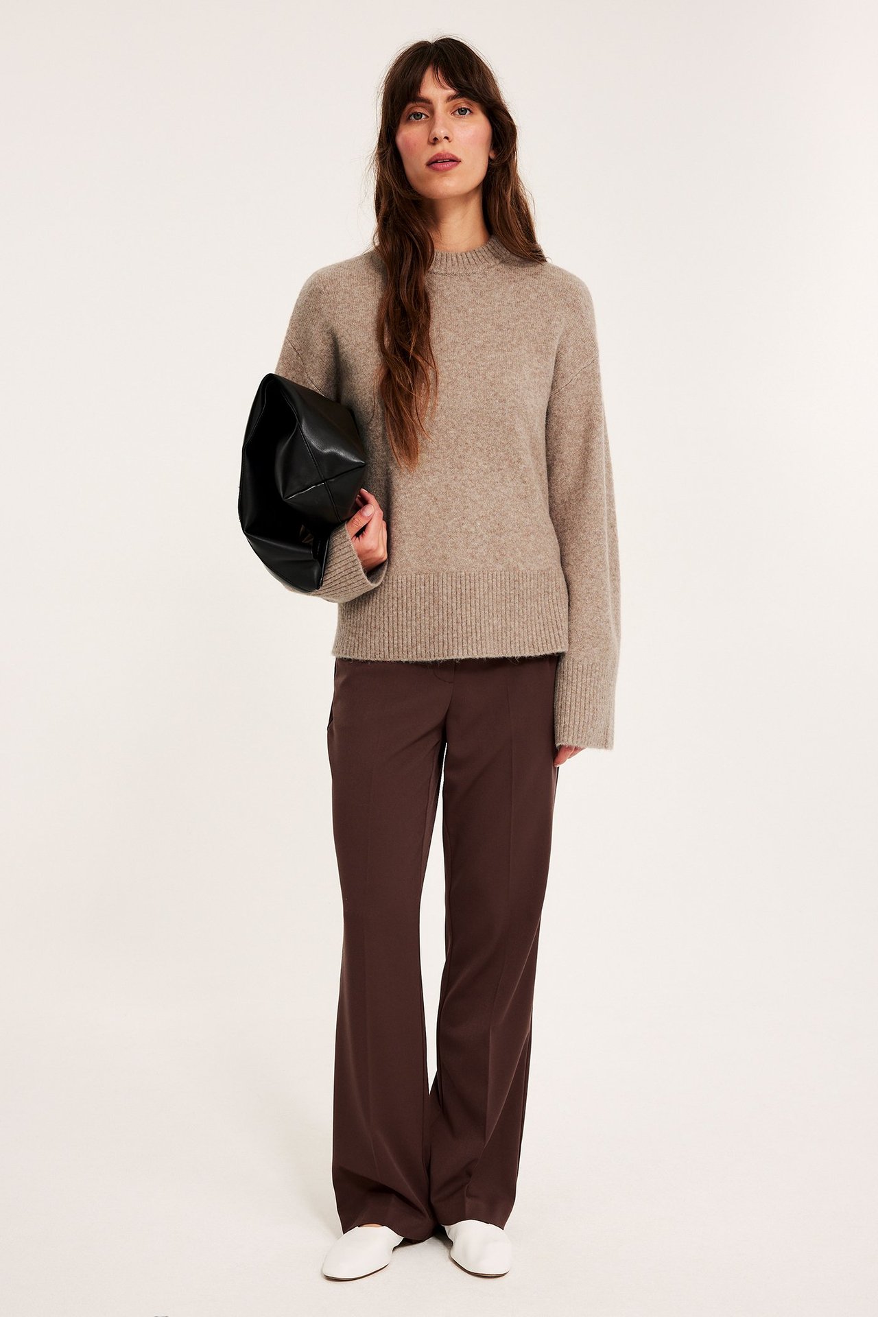 Edestä: nainen ruskeanbeigessä neuleessa, ruskeissa leveälahkeisissa housuissa, valkoisissa kengissä, pitäen mustaa laukkua.