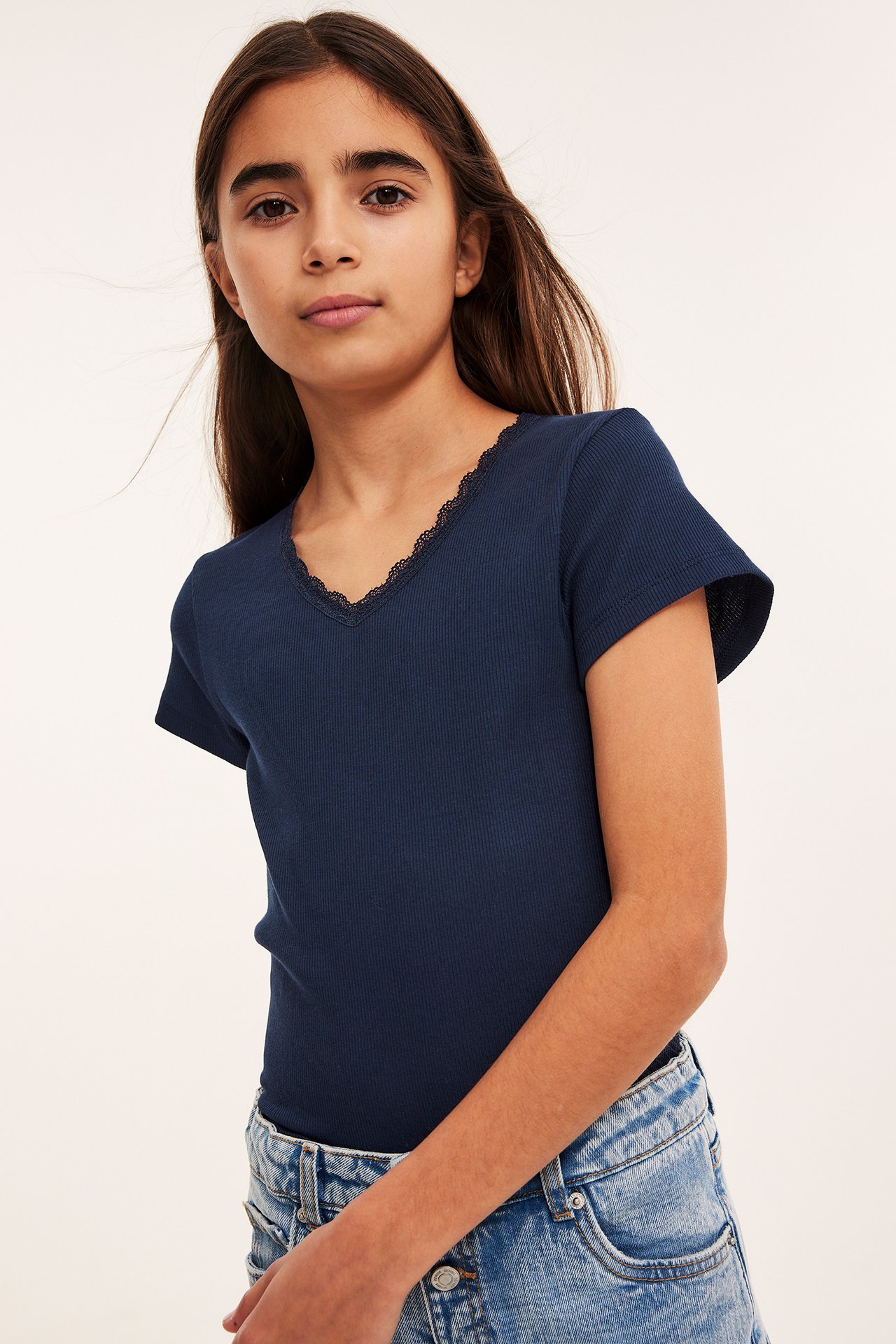 Framsida av ett barn i en åtsittande blå ribbad topp med svart spets och ljusblå jeans.