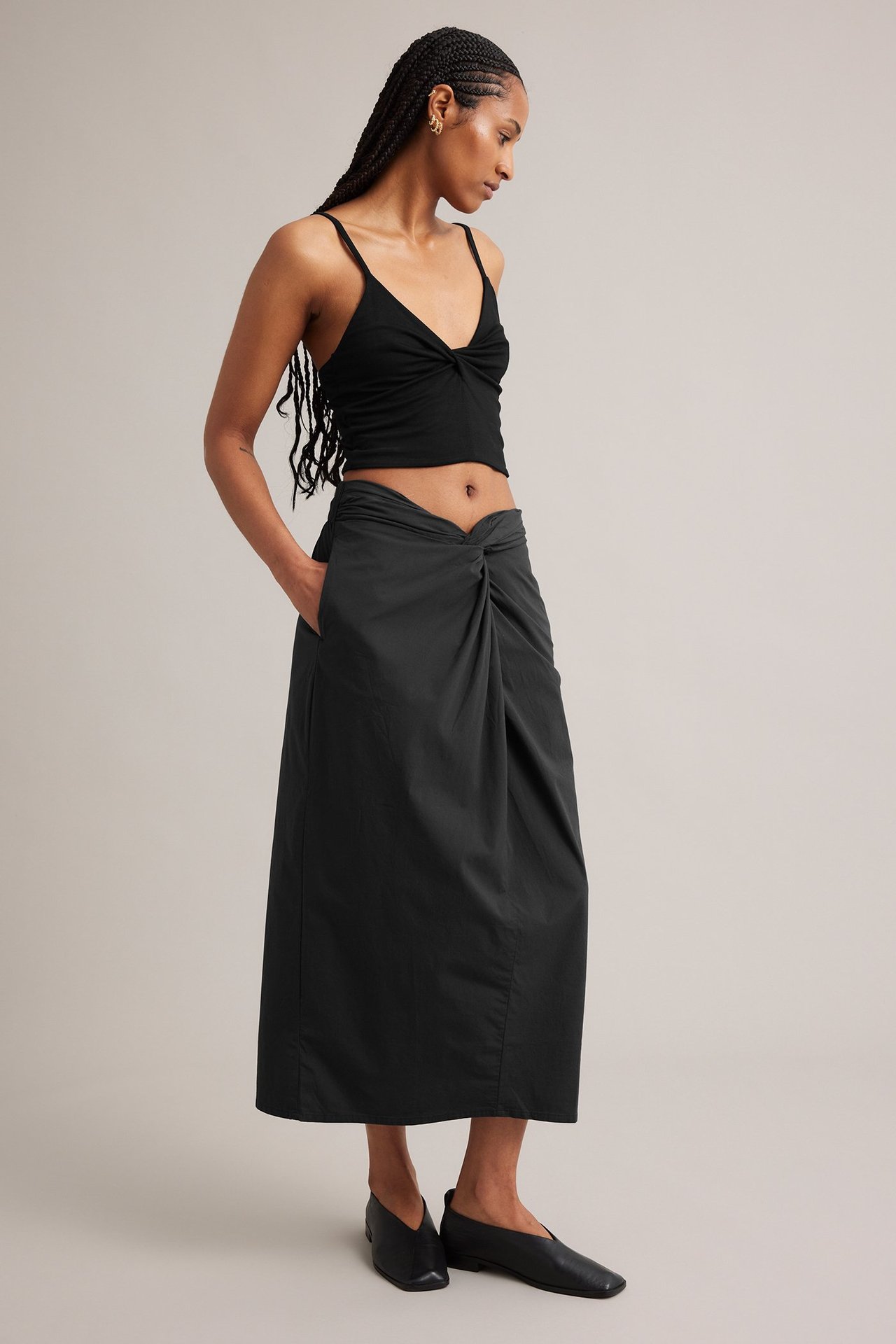 Sidebilde av en kvinne i svart midiskjørt med knutedetalj foran, svart camisole og svarte flate sko.