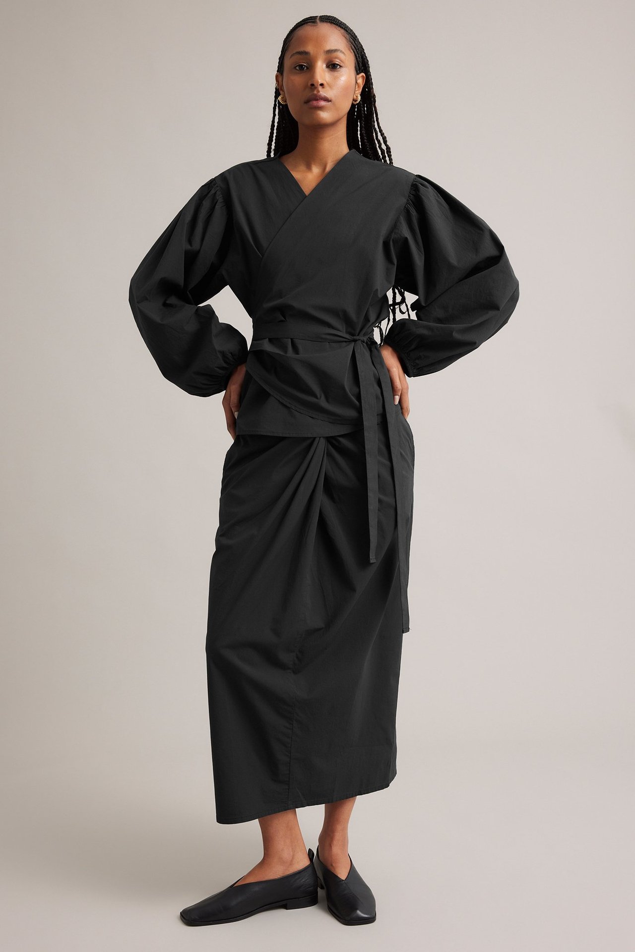Frontvisning: Kvinne i svart midiskjørt med knutedetalj, svart omslagstopp og svarte flate sko.