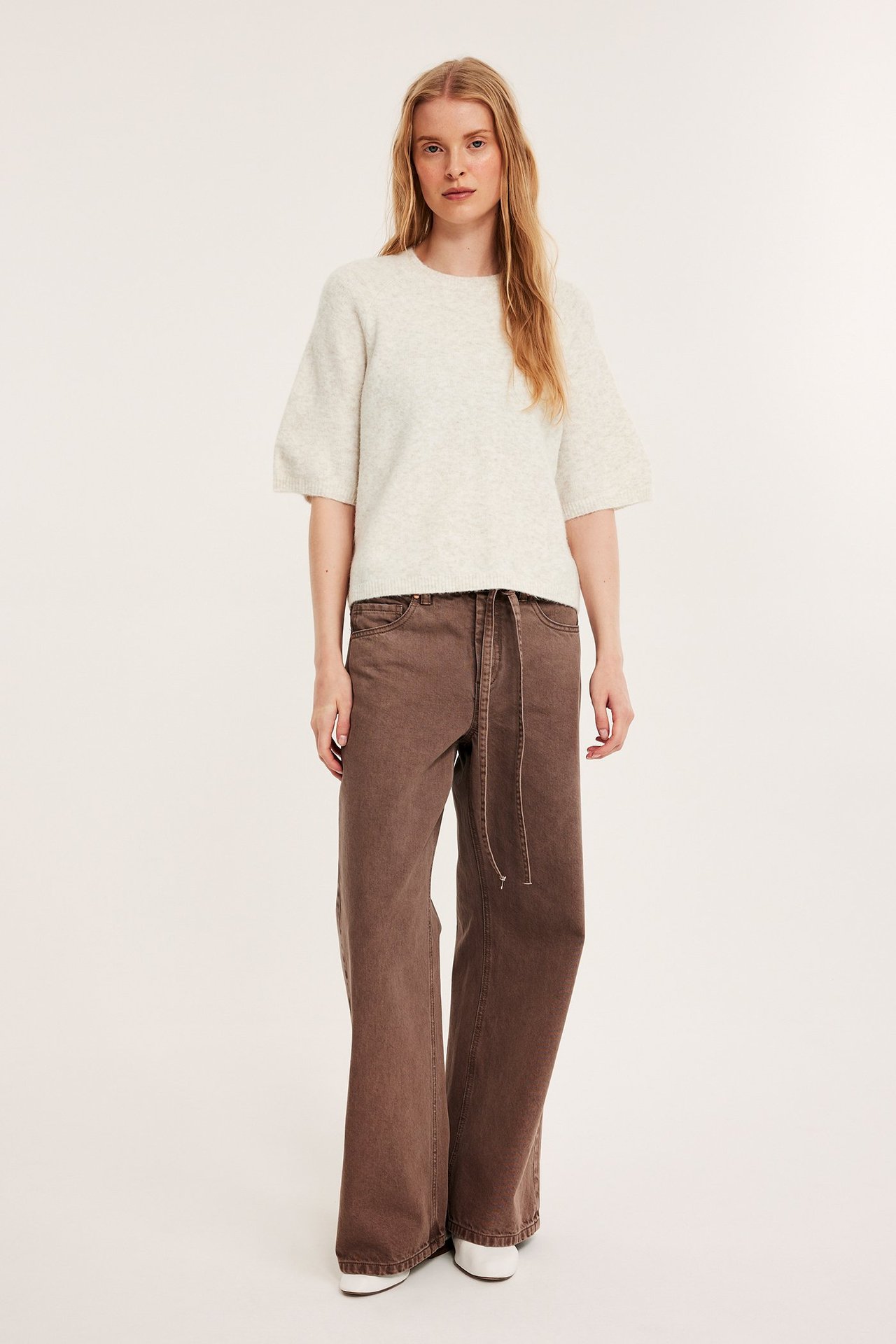 Framifrån: Kvinna i ljusbeige melerad oversized stickad t-shirt med voluminösa ärmar och bruna vida byxor.