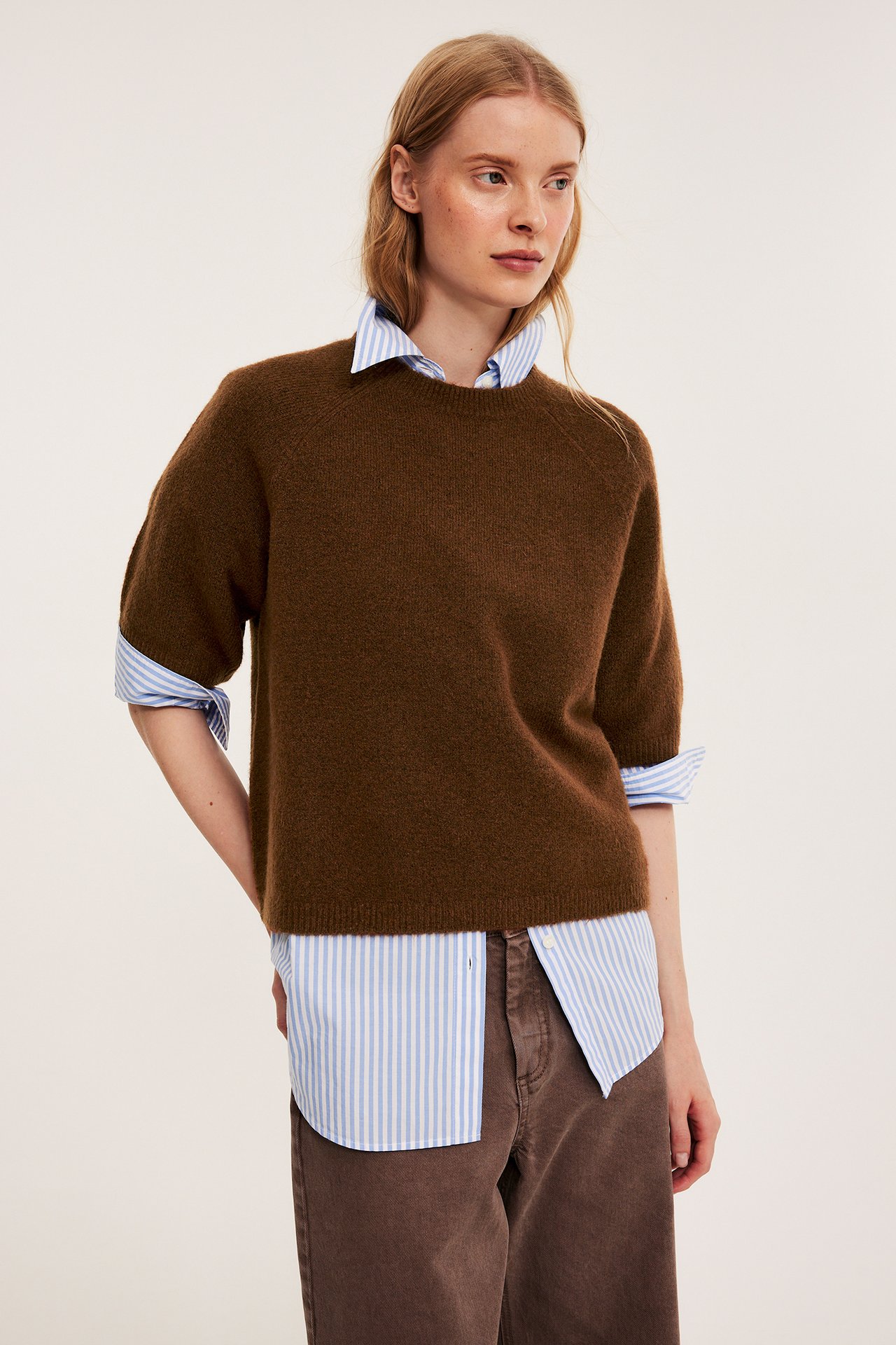 Frontvy av en kvinna i en oversized brun stickad t-shirt över en blårandig skjorta och bruna byxor.