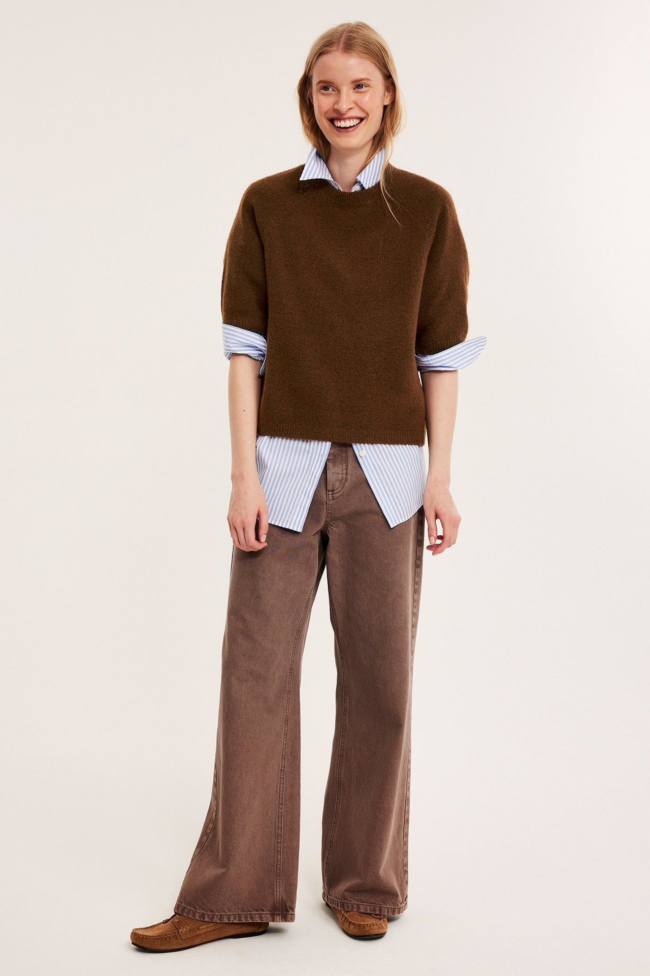 Kvinna bär en oversized brun stickad t-shirt över en randig skjorta, bruna vida byxor och mockasiner.