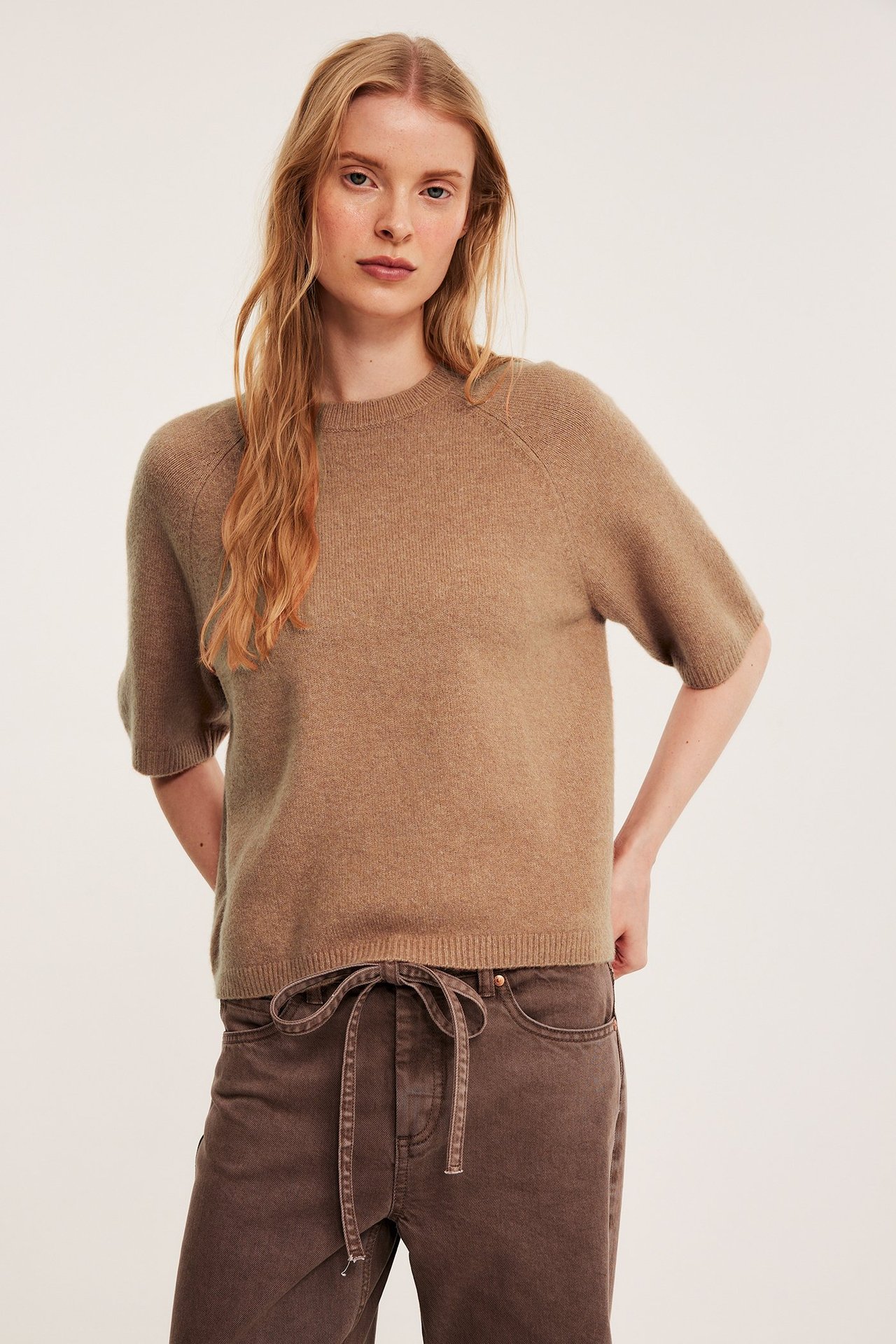 Framifrån, kvinna bär en oversized ljusbrun melerad stickad kortärmad topp och mörkbruna byxor.