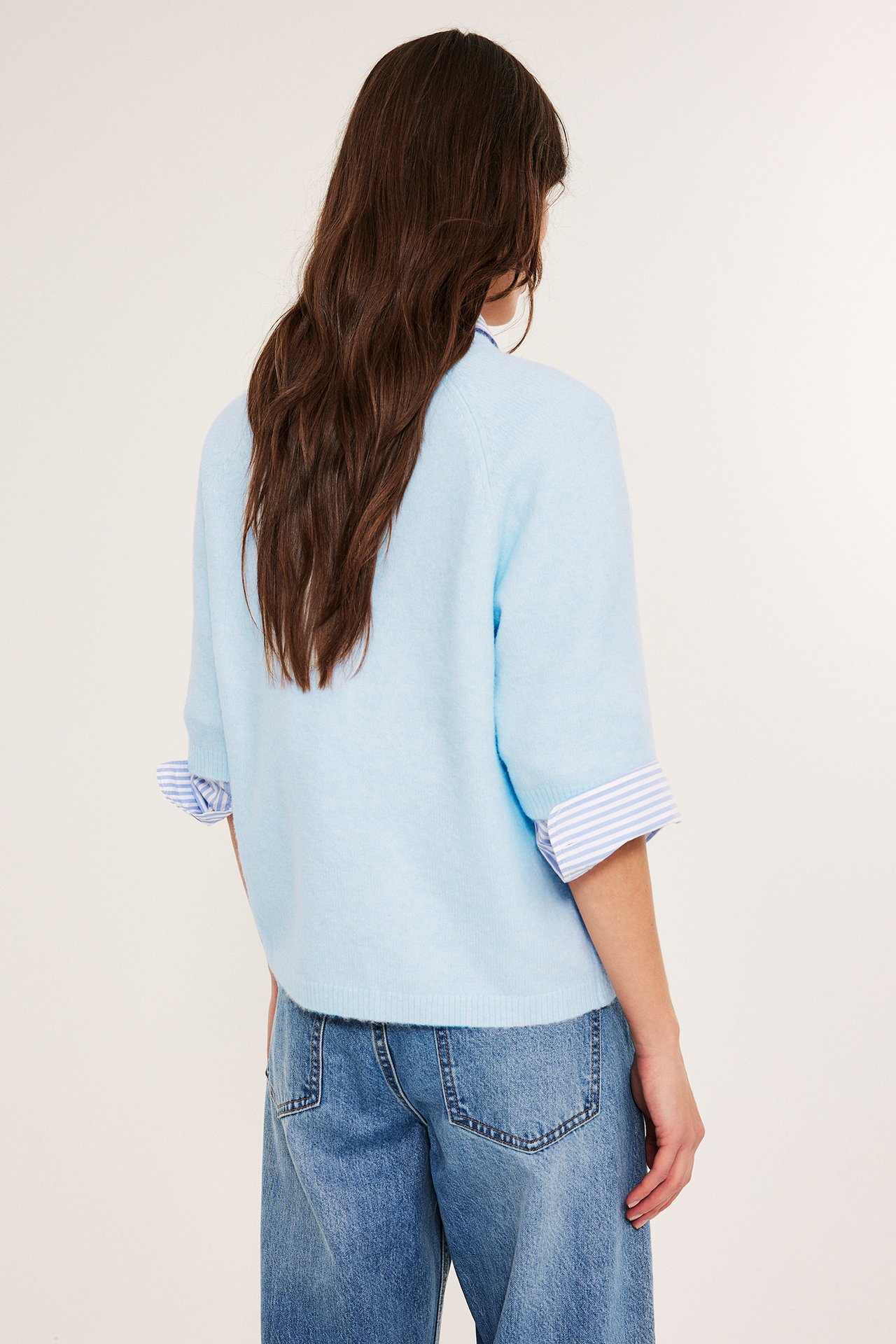 Widok z tyłu kobiety w jasnoniebieskim, oversize'owym swetrze, koszuli w paski i niebieskich jeansach.
