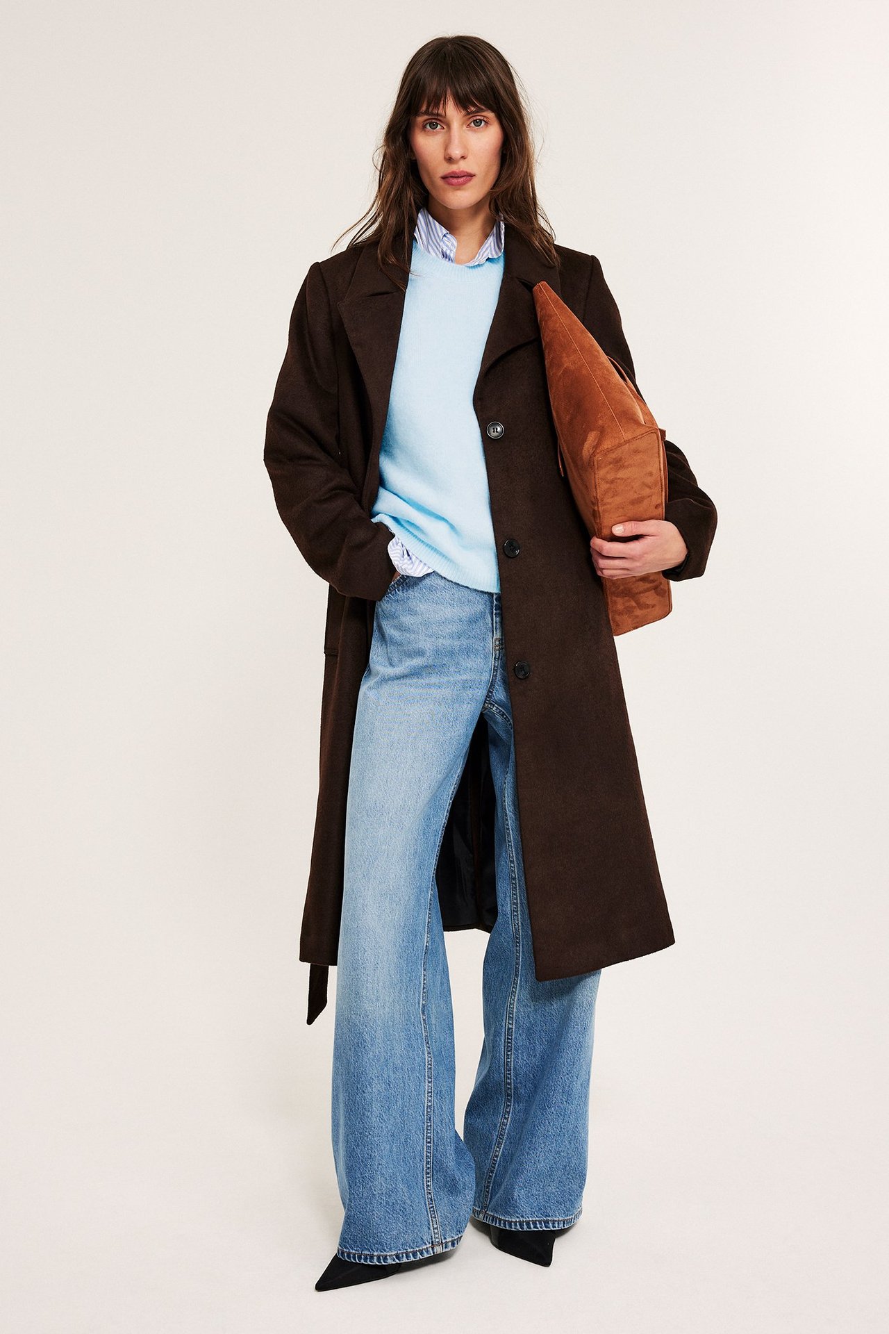 Frontvy: Kvinna i ljusblå oversized stickad tröja, brun kappa, ljusa jeans, håller en brun väska.