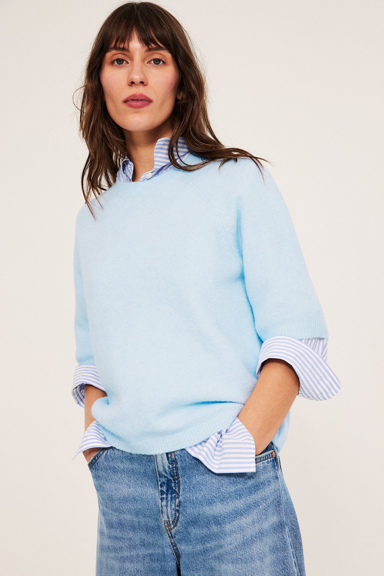 Widok z przodu kobiety w jasnoniebieskim oversizowym dzianinowym T-shirt-swetrze, koszuli w paski i niebieskich jeansach.