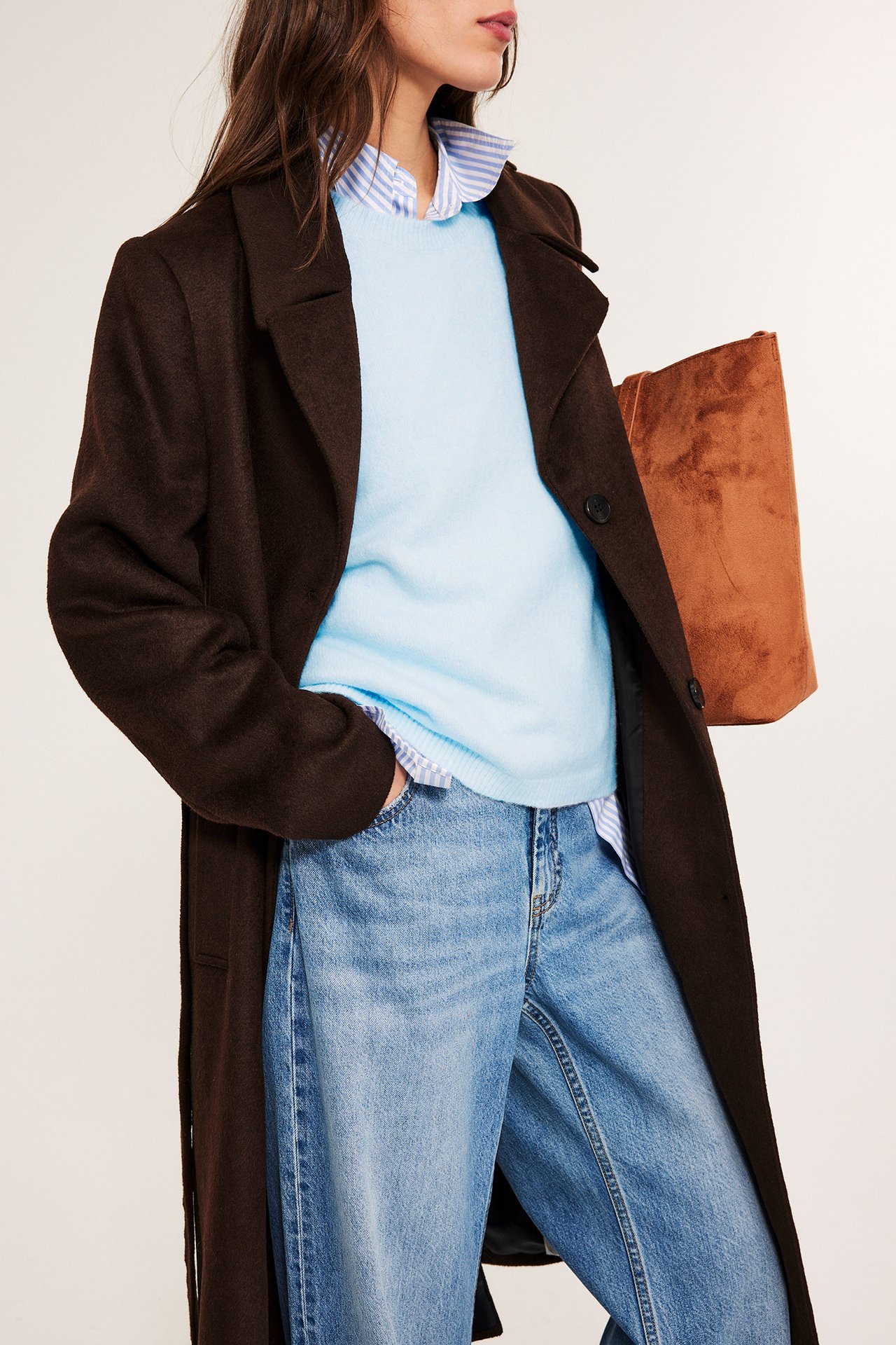 Framifrån: Kvinna i ljusblå oversized stickad t-shirt, randig skjorta, brun kappa, jeans, håller brun tygkasse.