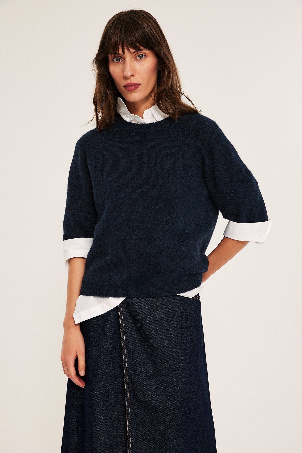 Front view: Kvinna i mörkblå oversized stickad t-shirt över vit skjorta, mörkblå denim omlottkjol.