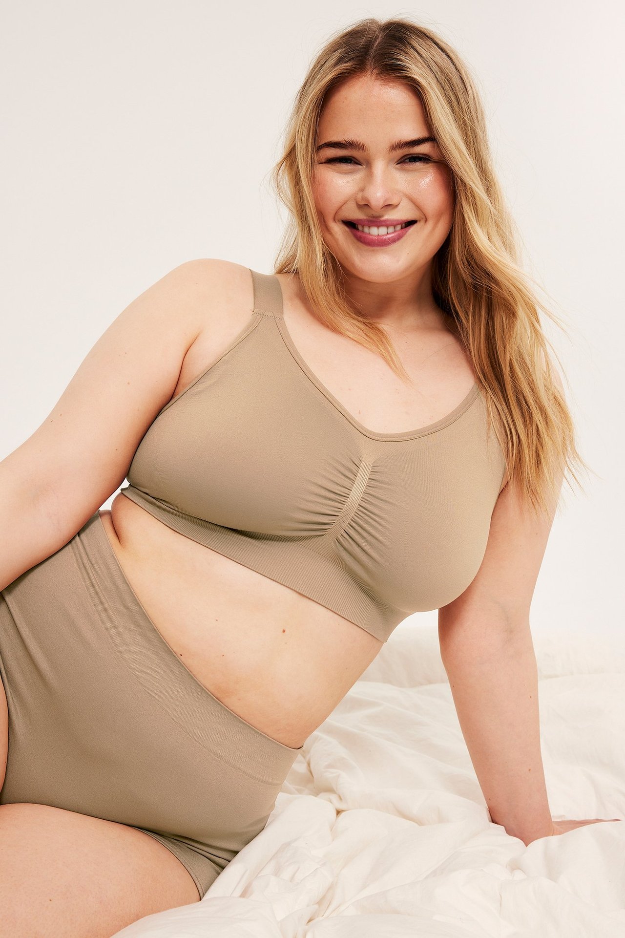 Framifrån: leende kvinna i beige sömlös bygellös bh med rynkning och matchande högmidjade trosor.
