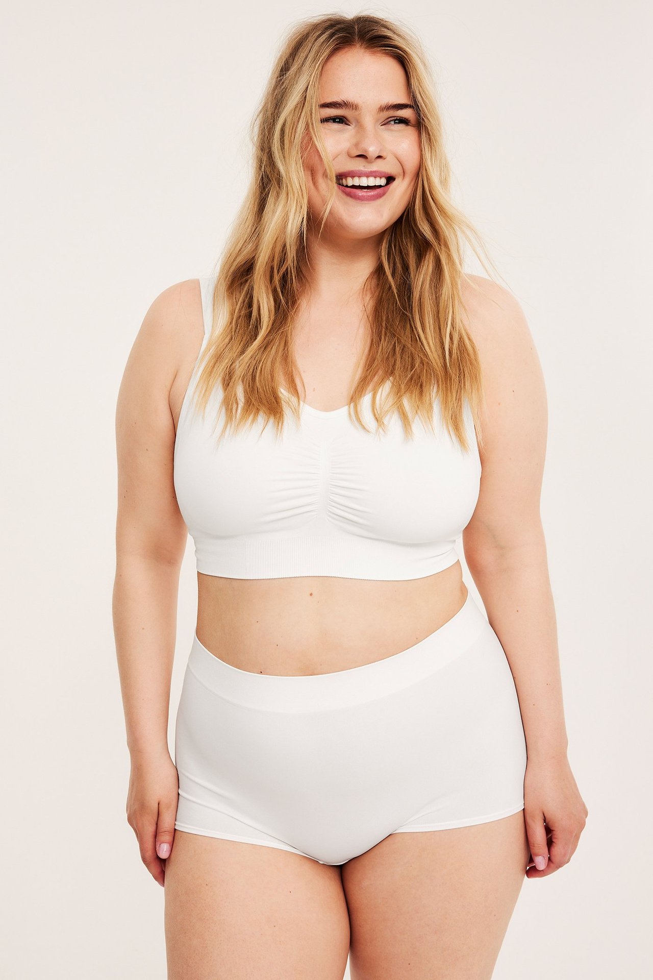 Frontbilde av en smilende kvinne i hvit sømløs boksertruse og matchende hvit sømløs bralette.