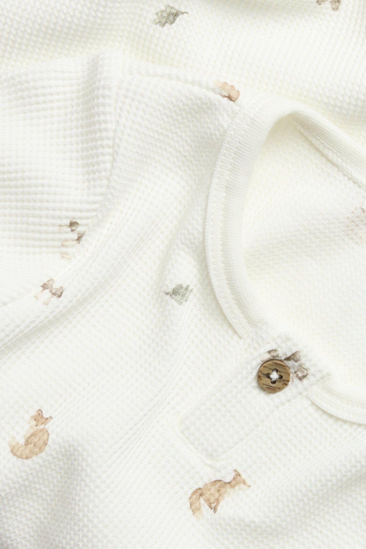 Nærbilde av en offwhite vaffelstrikket babybody med skogsdyrmønster og en treknapp.