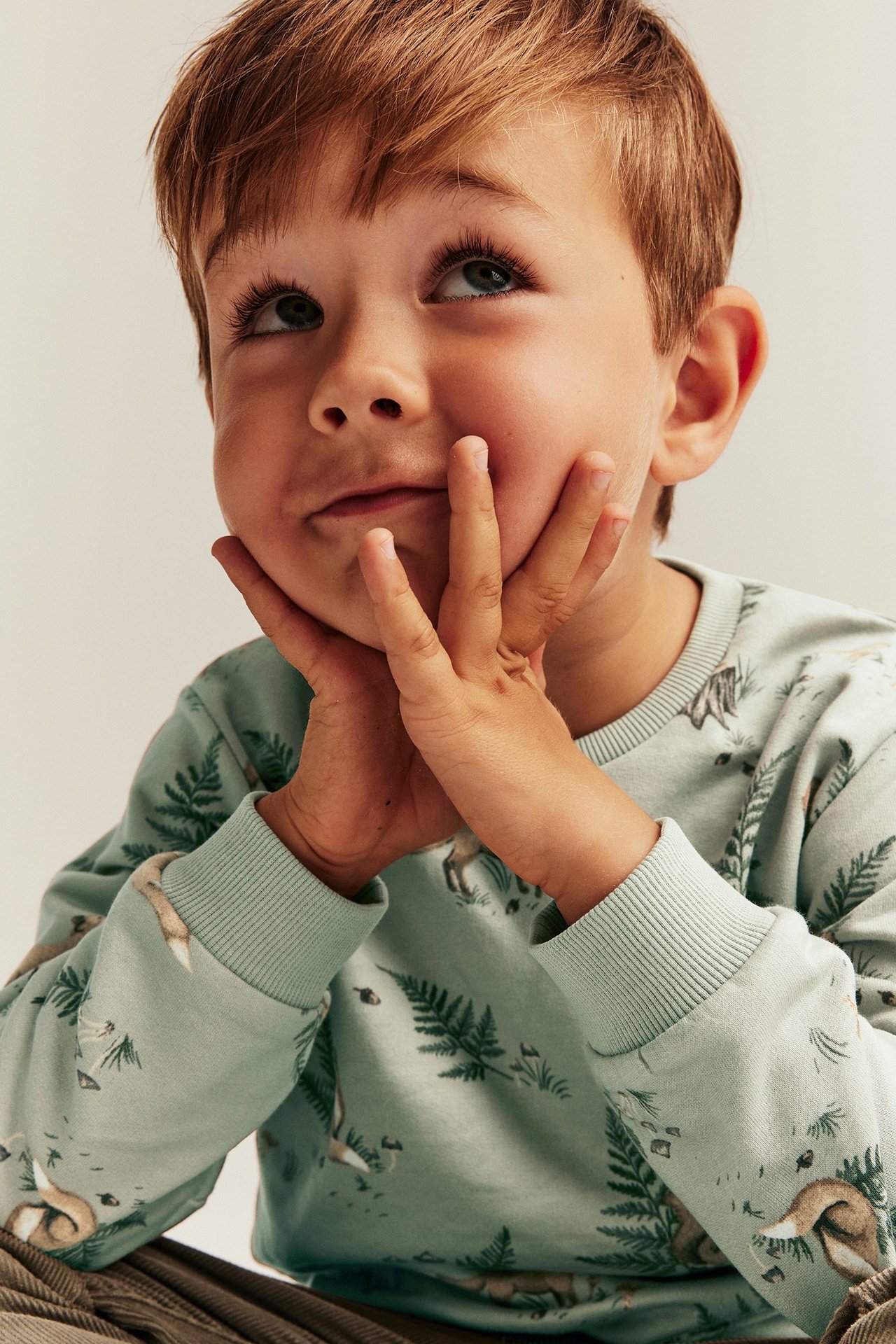 Frontalvisning, barn i en lys grønn bomullsgenser med skogsmønster, hendene på kinnene.
