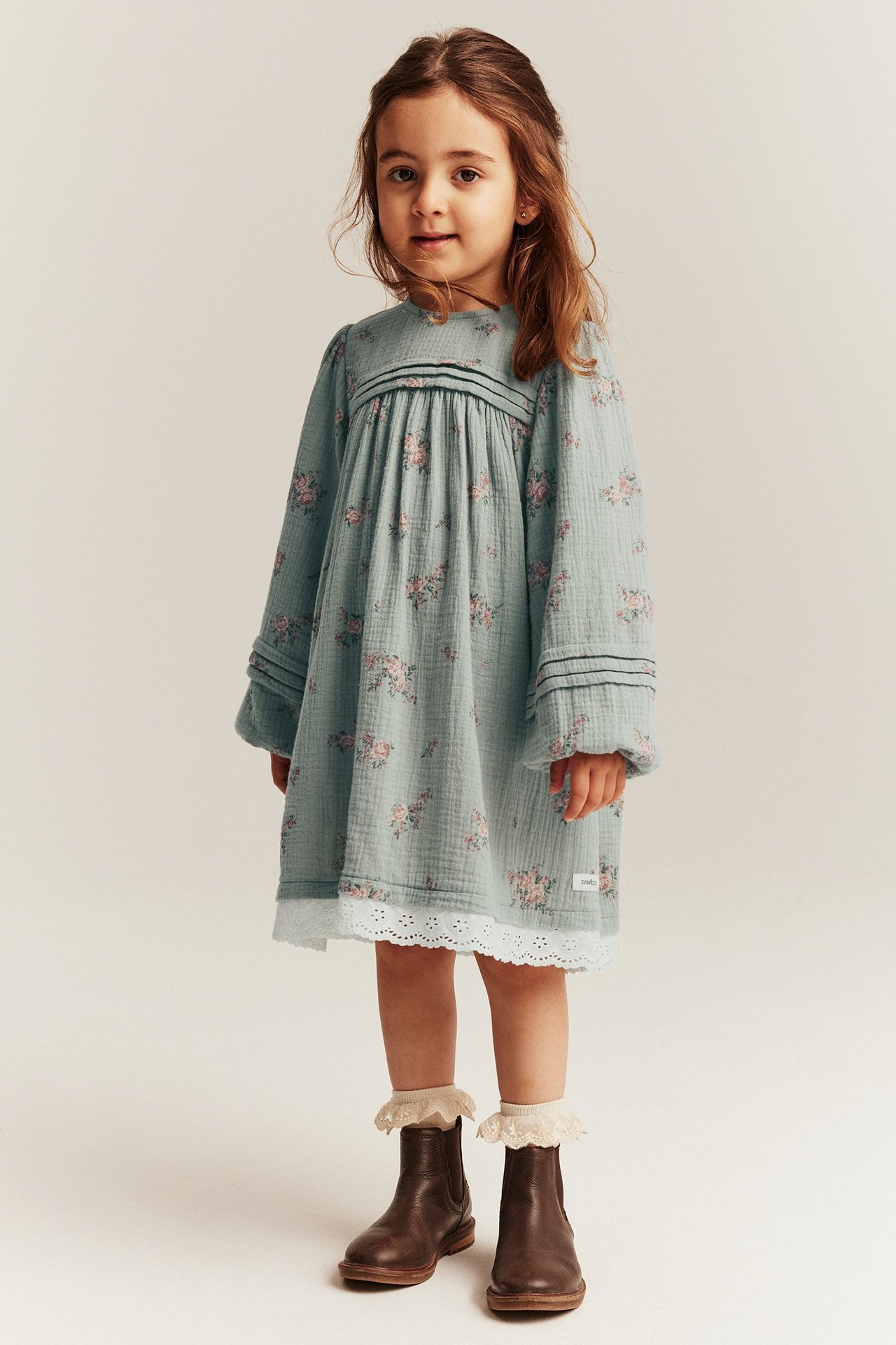 Widok z przodu: dziecko w jasnozielonej sukience w kwiaty z bawełny, z zakładkami, koronką, kremowymi skarpetkami, brązowymi butami.