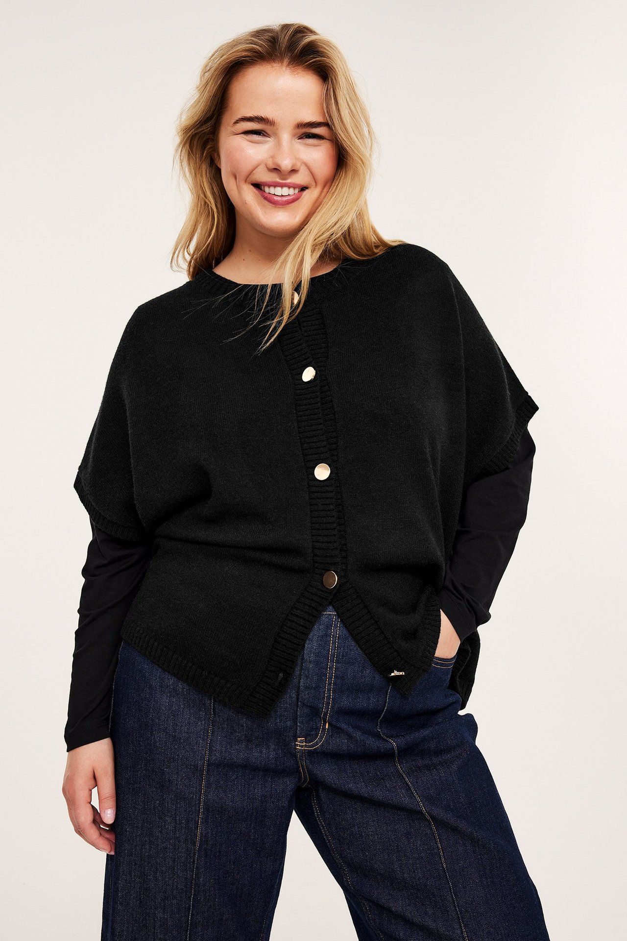 Framifrån: kvinna i svart stickad väst med guldknappar, svart topp och mörkblå jeans.