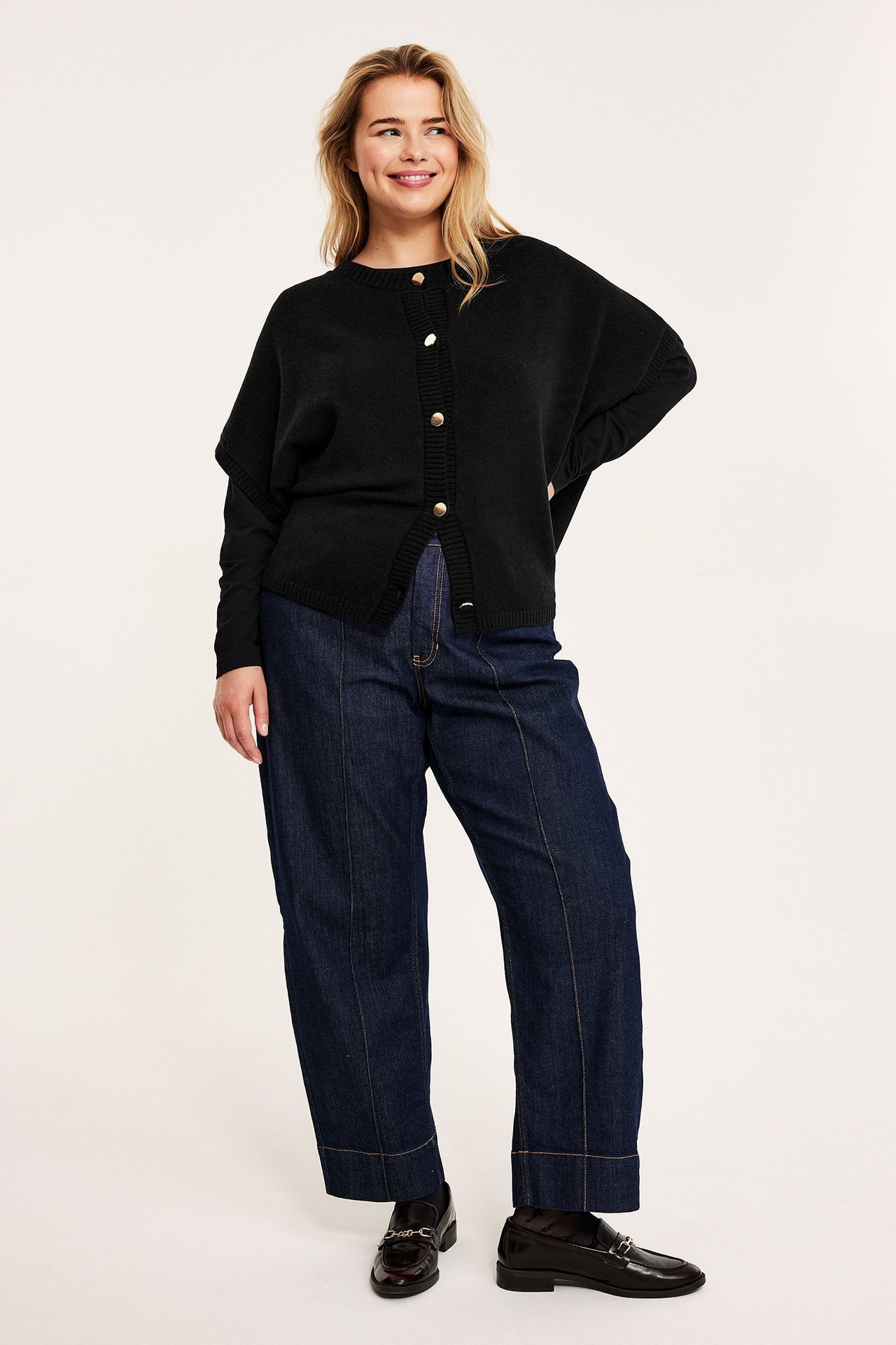 Framifrån: Kvinna bär svart stickad väst med guldfärgade knappar, svart topp, mörkblå jeans, bruna loafers.