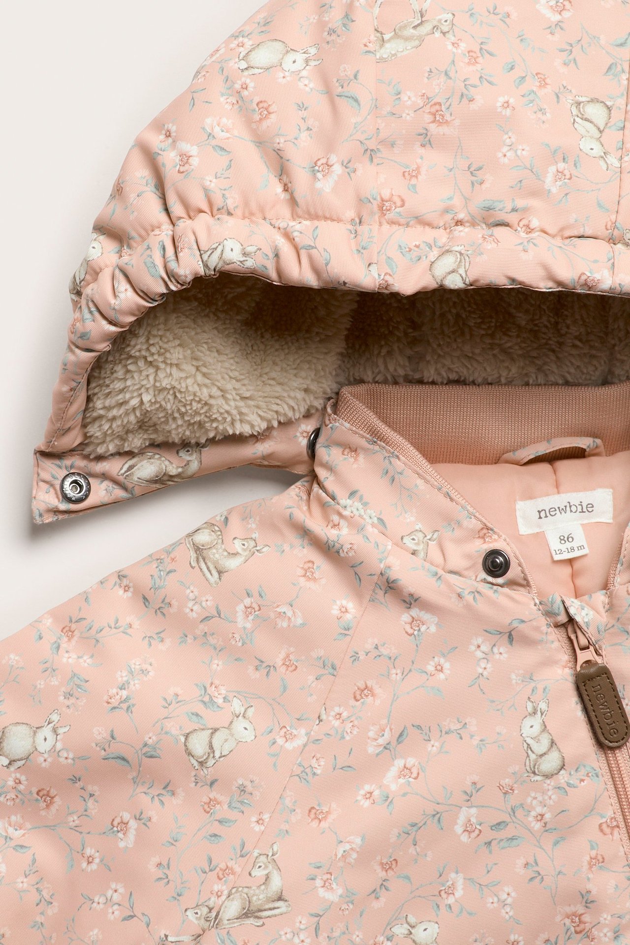 Nærbilde av en rosa babyoverall med blomster- og dyremotiv, med fluffy fôret hette og glidelås.