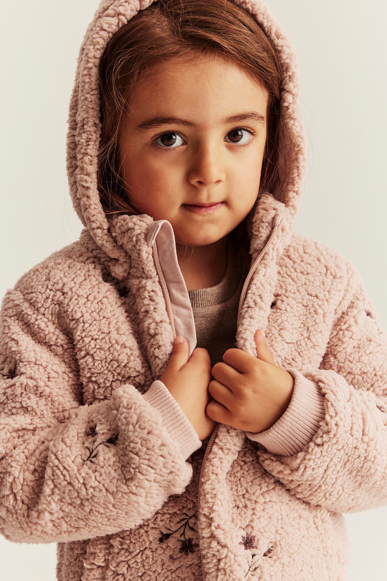 Nærbilde forfra av et barn i en rosa hettejakke i pile med blomsterbroderi og glidelås.