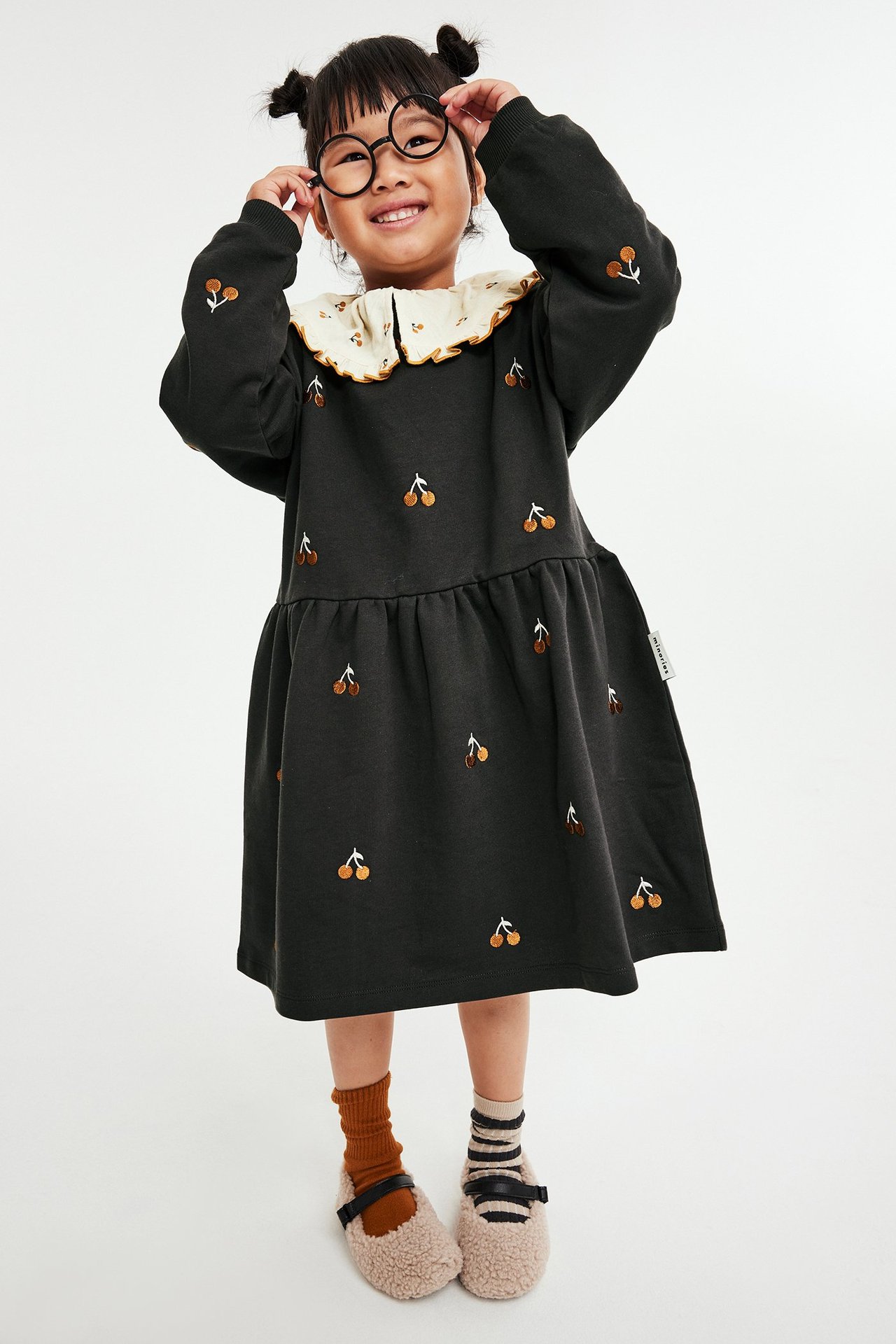 Widok z przodu: Dziecko w czarnej sukience z długim rękawem, haftem wiśni, falbankowym kołnierzem, okularach i futrzanych butach.
