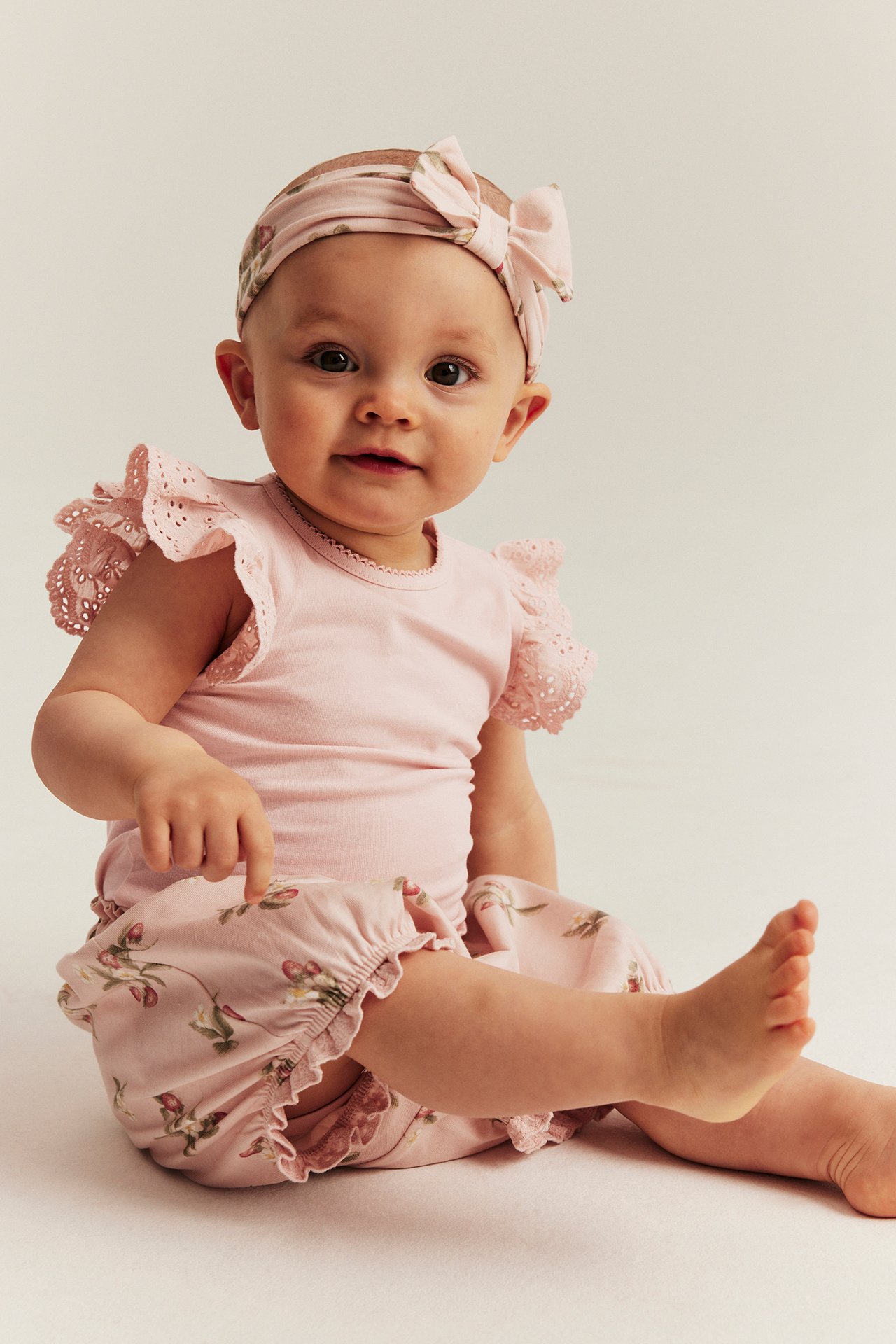 Frontvisning av en baby i lyserosa bomullsbody med blondevolangermer, blomstrete bloomers og matchende sløyfebånd.