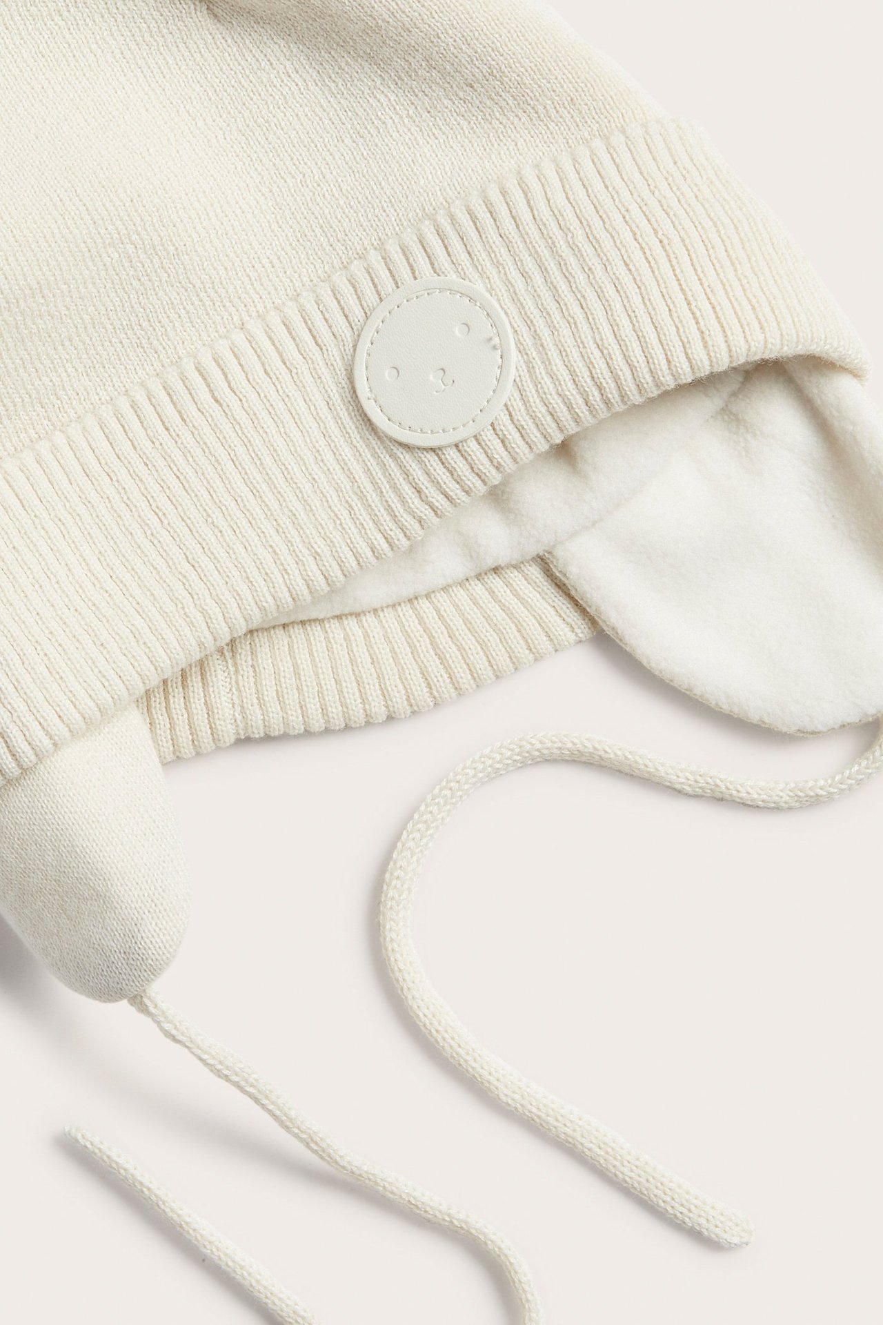 Jasnobeżowa dzianinowa czapka niemowlęca z nausznikami, wiązaniami, polarową podszewką i etykietą. Zbliżenie.