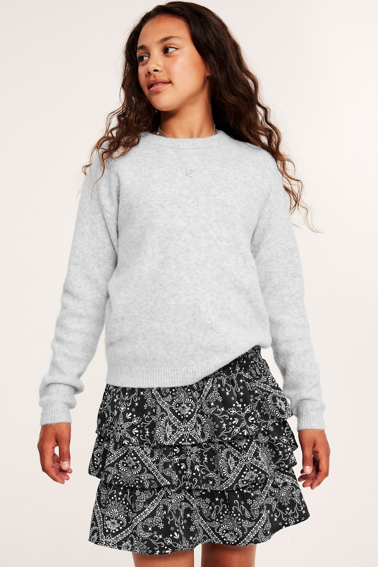 Framsida av ett barn i en ljusgrå melerad finstickad tröja och en svart mönstrad volangkjol.