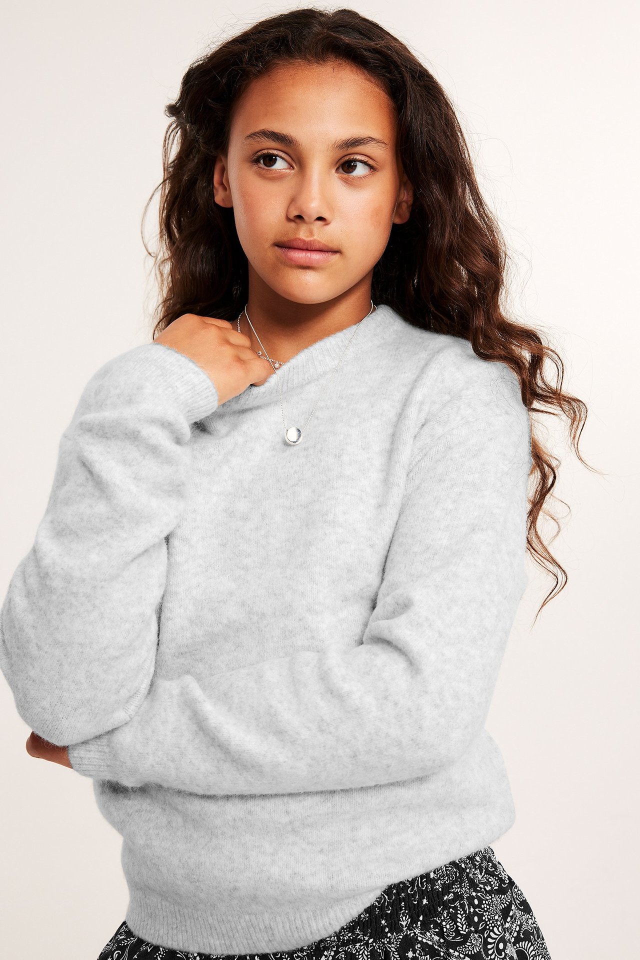 Widok z przodu: Dziecko w jasnoszarym melanżowym swetrze, srebrnym naszyjniku i czarnej wzorzystej spódnicy.