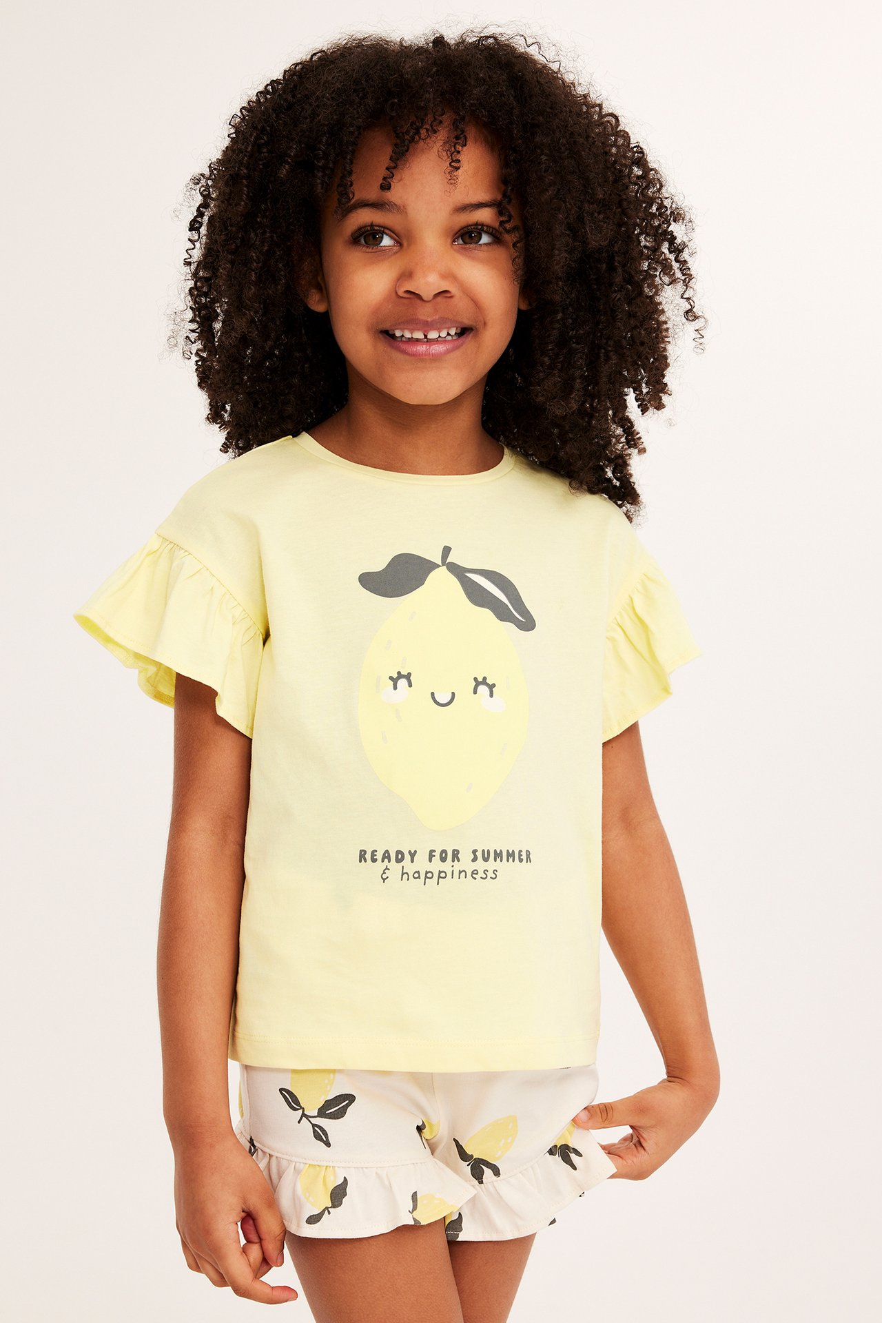 Frontvy: Barn i ljusgul volangärmad t-shirt med citrontryck och ljusbeige mönstrade shorts.