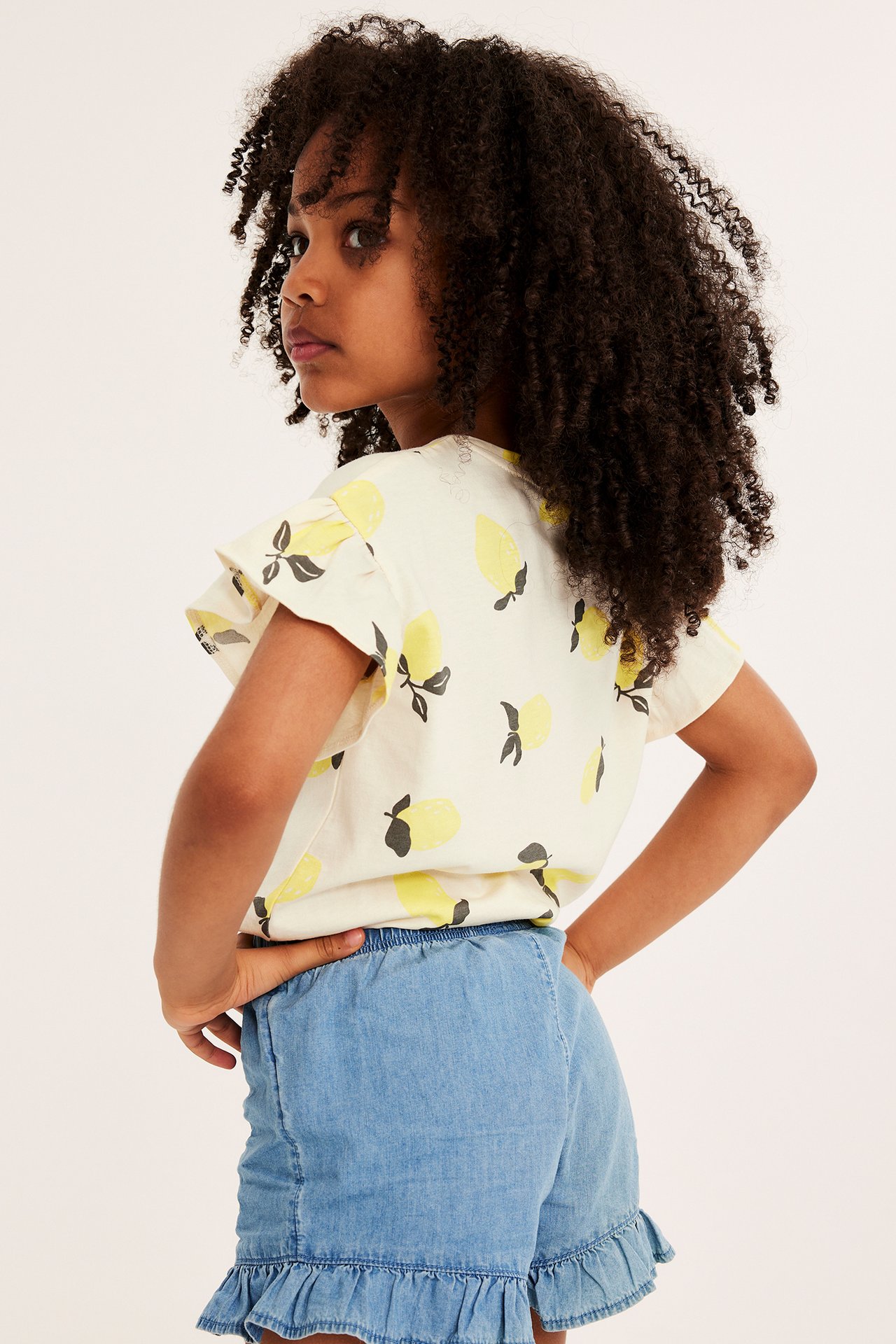 Barn, bakifrån: offwhite bomullstopp med volangärm, citronprint och ljusblå denimshorts.