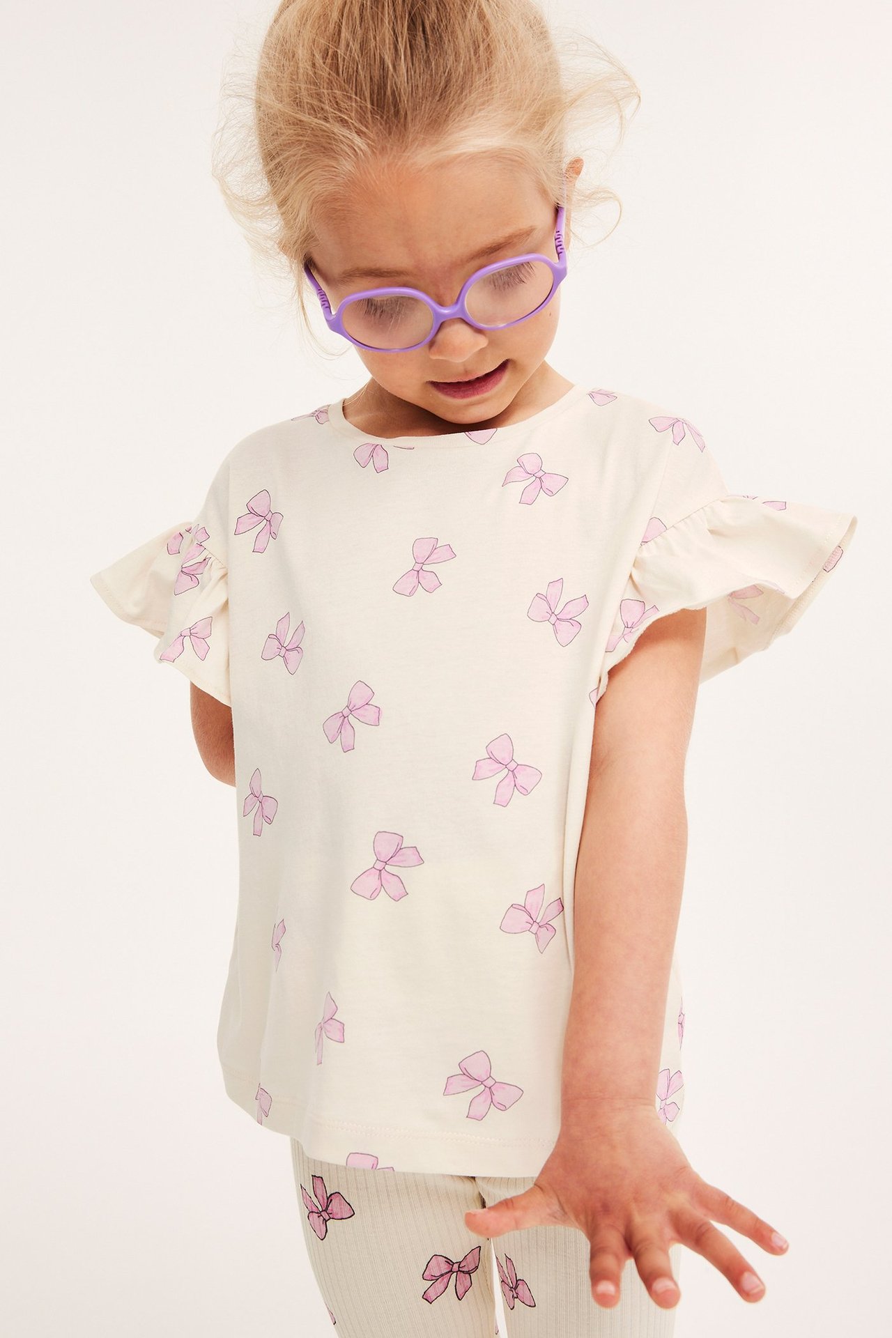 Frontvy av ett barn i en vit trikåtopp med volangärm, rosa rosettprint och matchande leggings.