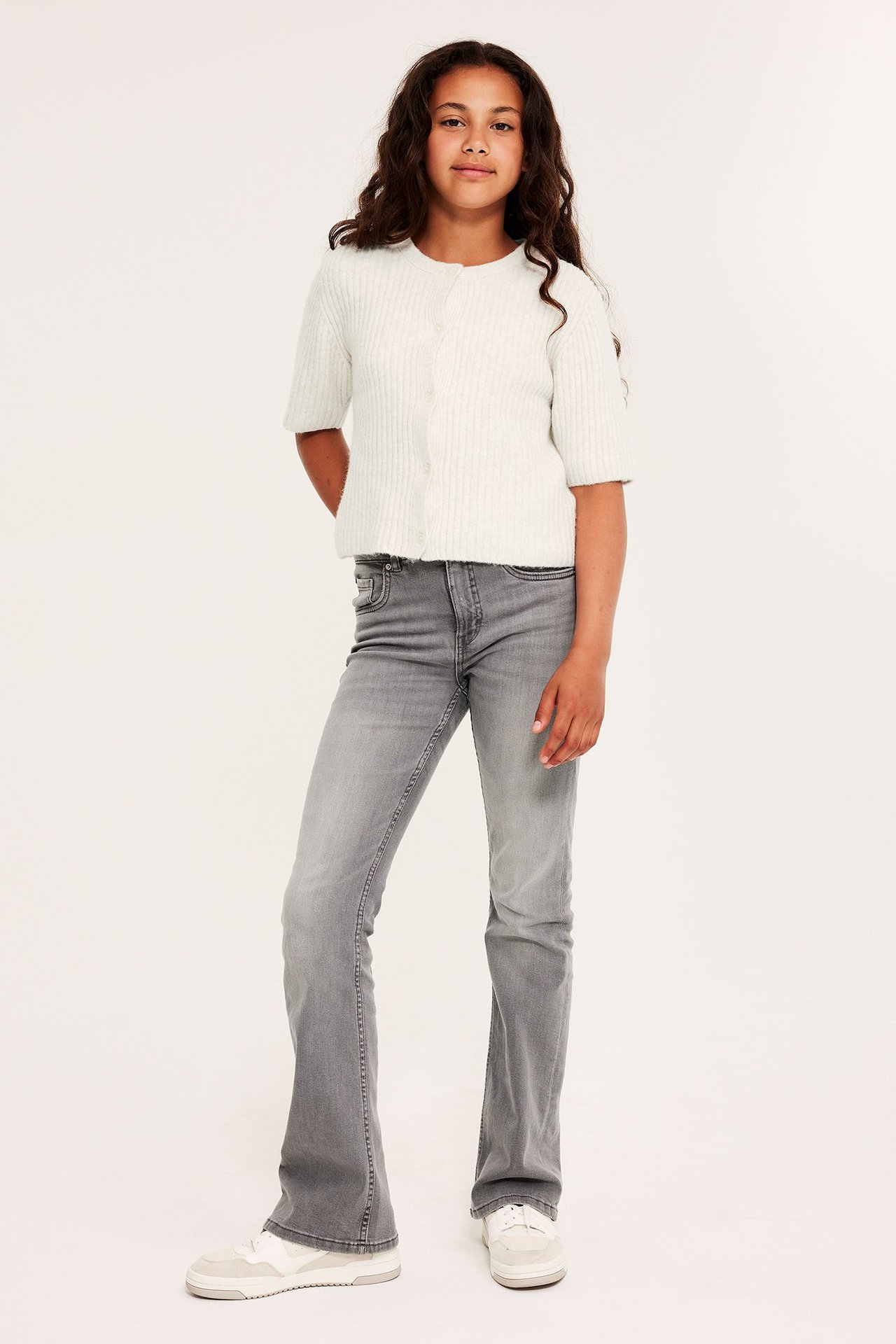 Frontbilde av et barn i en hvitmelert kortermet strikket cardigan, grå slengbukser og hvite joggesko.