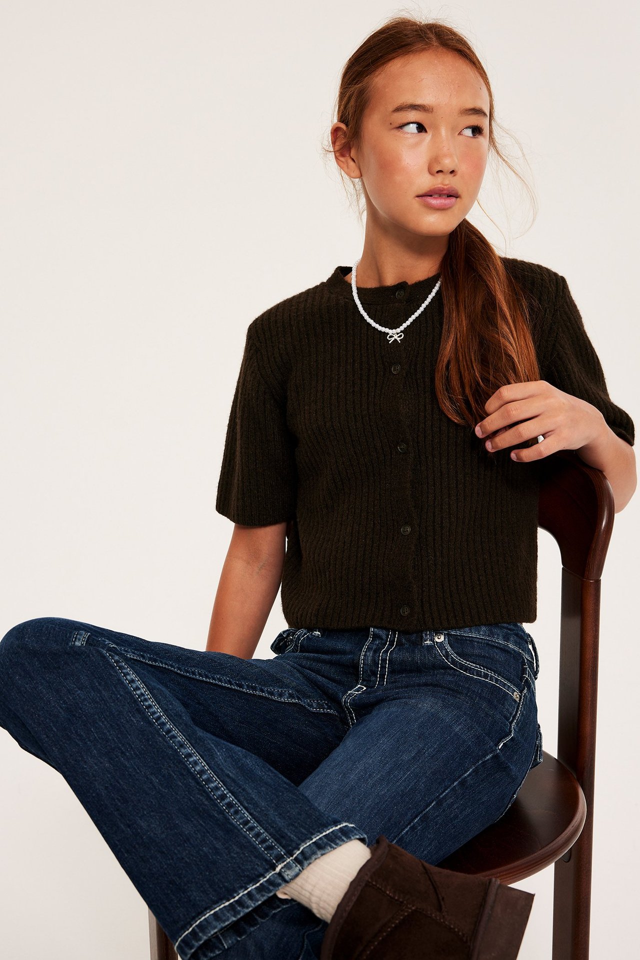 Framifrån, ett barn i mörkbrun ribbad kortärmad kofta, mörkblå jeans, pärlhalsband och bruna stövlar.