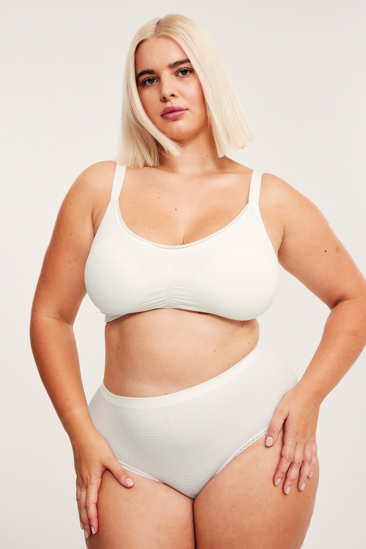 Forfra, kvinne i hvit sømløs strukturert hipstertruse med blonde og matchende BH.