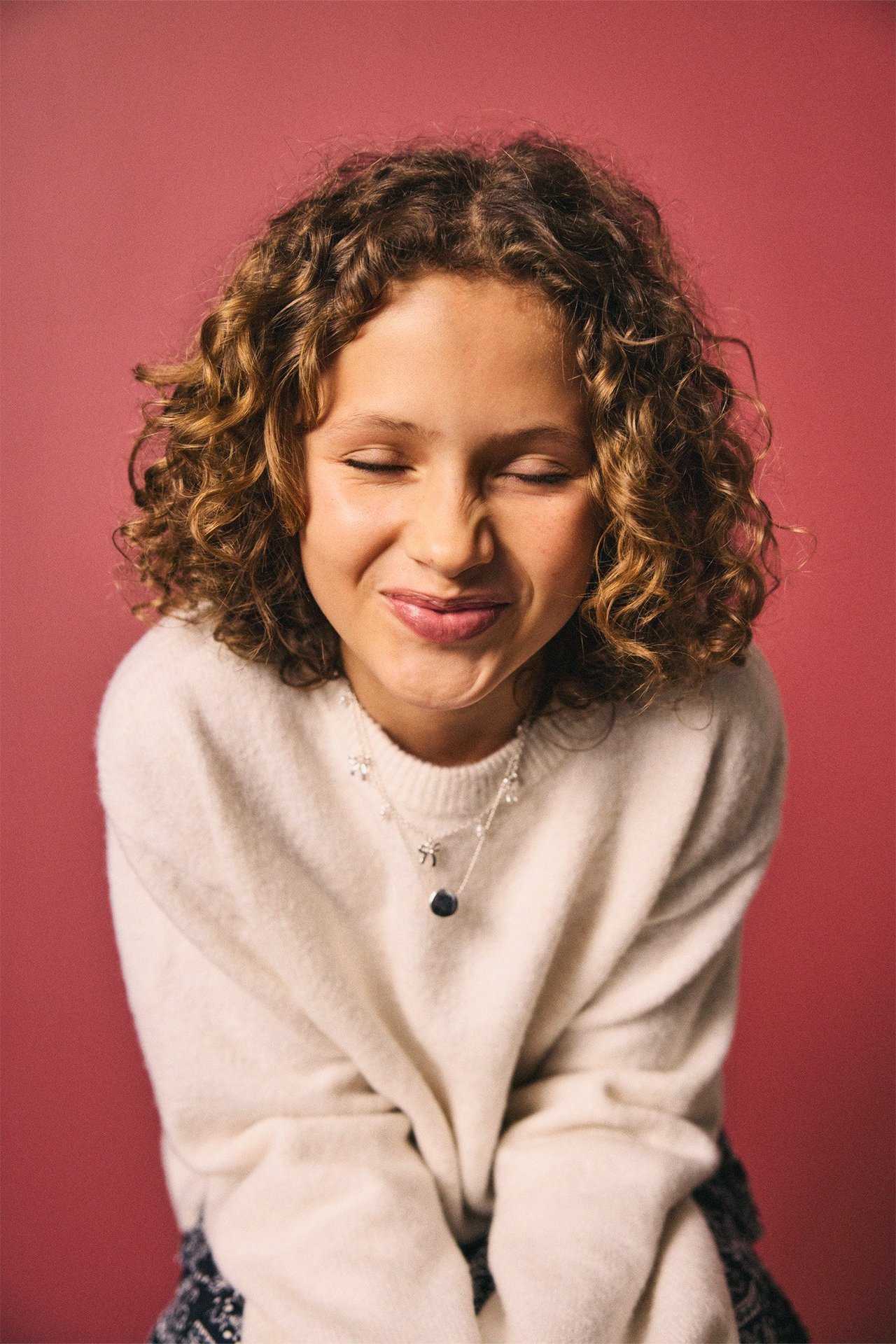 Edestä kuvattu lapsi yllään luonnonvalkoinen meleerattu neulepusero ja kerroksellinen kaulakoru.