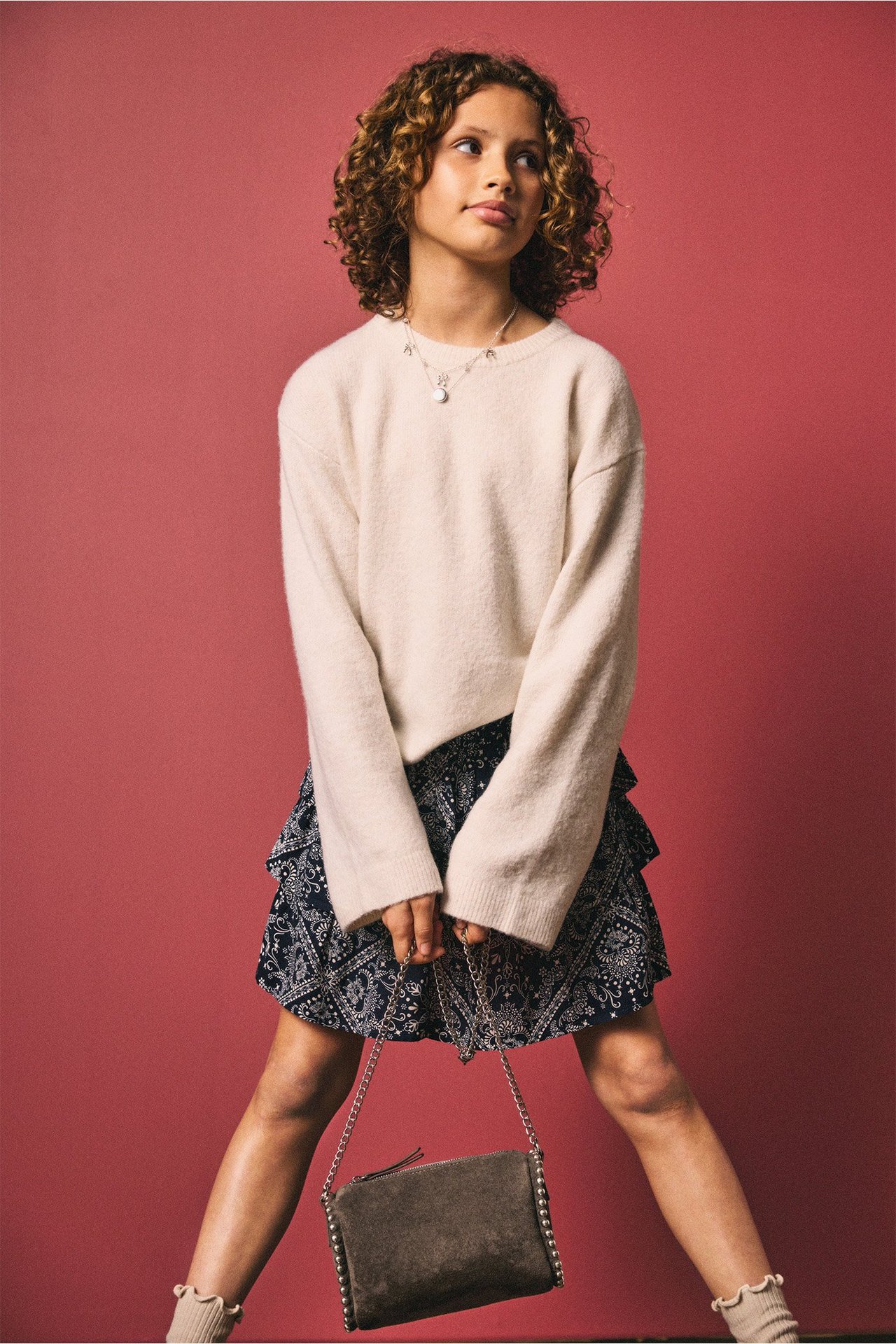 Dziecko w białym melanżowym swetrze, granatowej spódnicy we wzory, srebrnym naszyjniku, trzyma szarą torebkę z nitami.