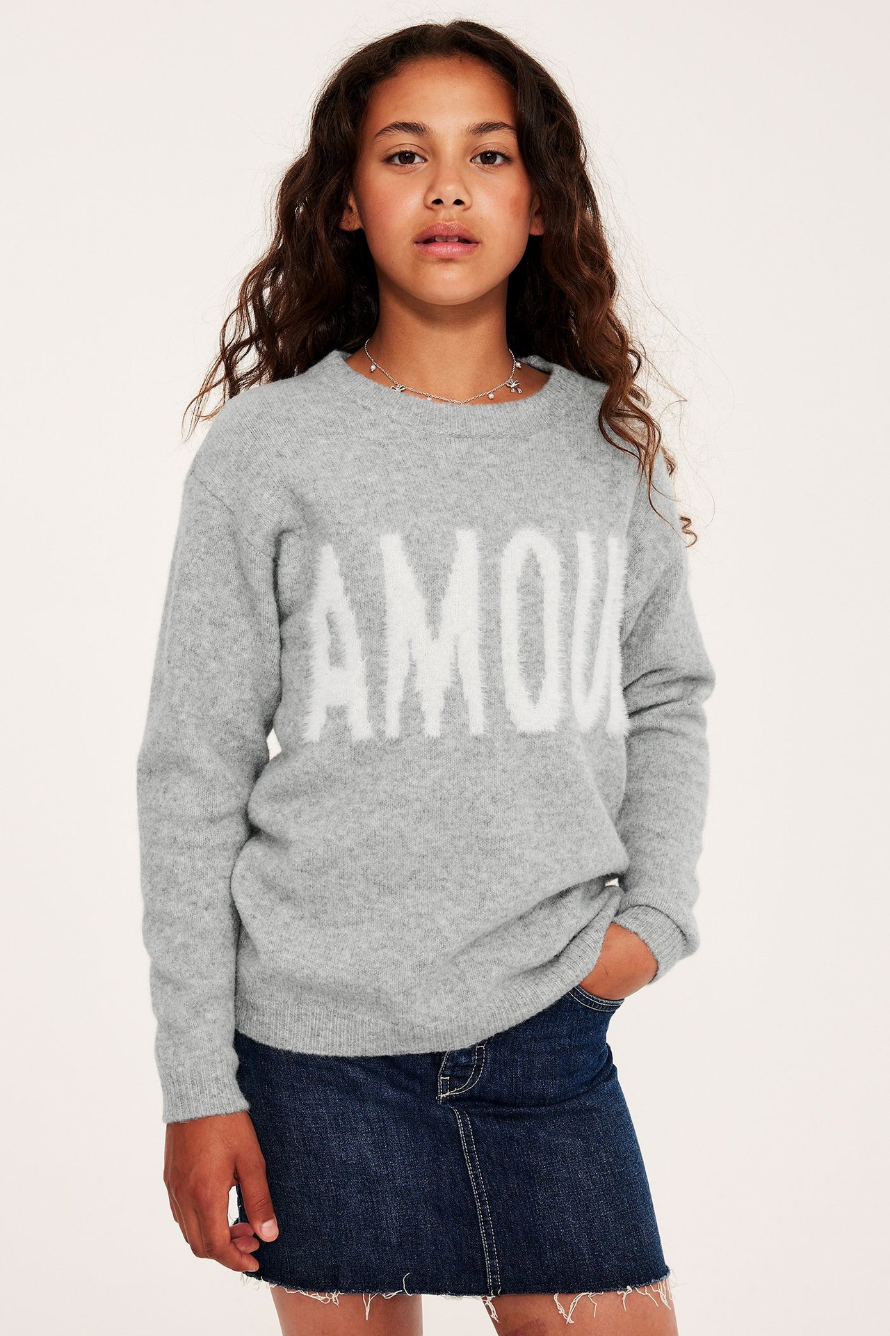 Frontvy: Barn i gråmelerad stickad tröja med vit 'AMOUR'-text, mörkblå jeanskjol, silverhalsband.