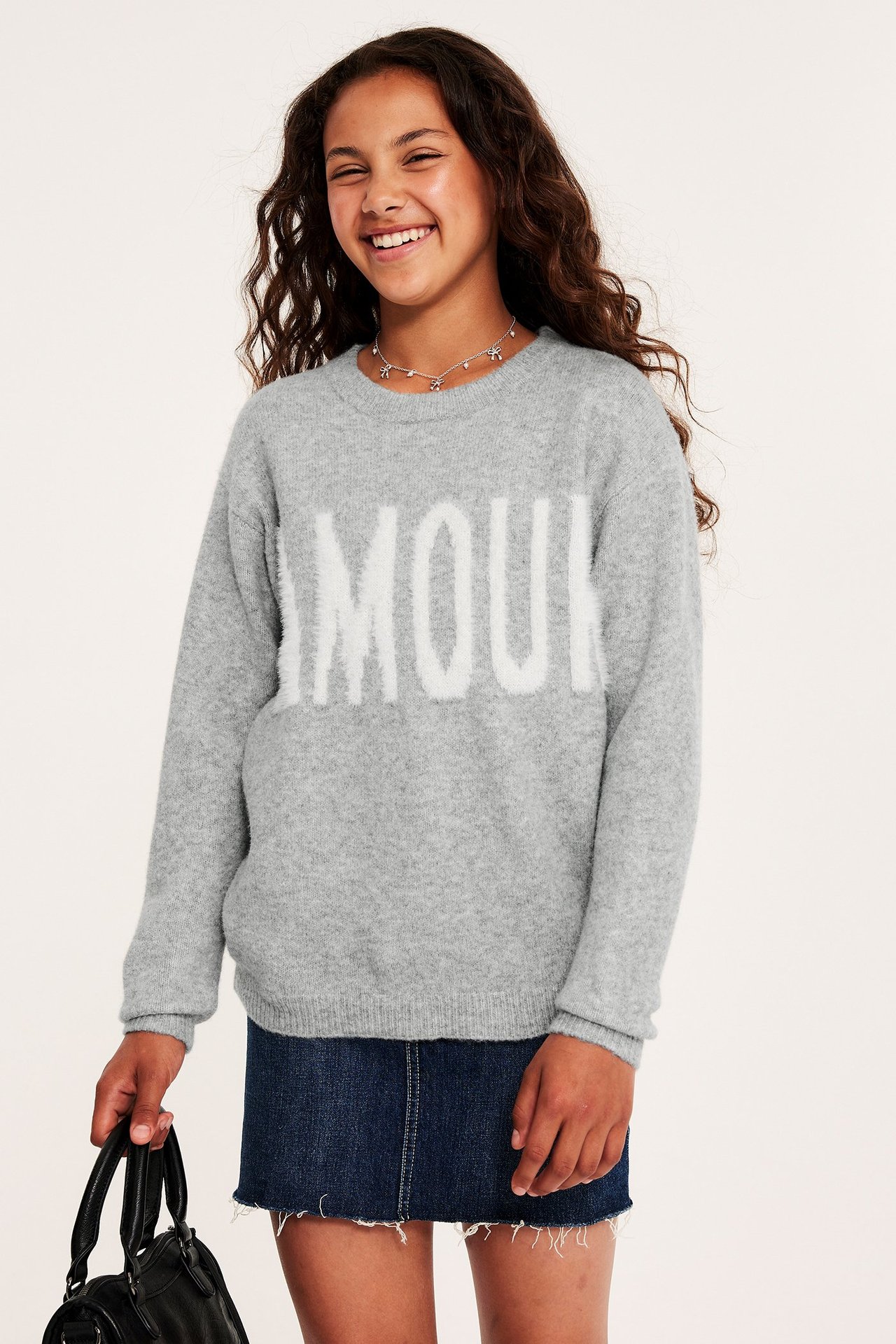 Widok z przodu: Dziecko w szarym melanżowym swetrze "AMOUR", granatowej spódnicy jeansowej, trzyma czarną torebkę.