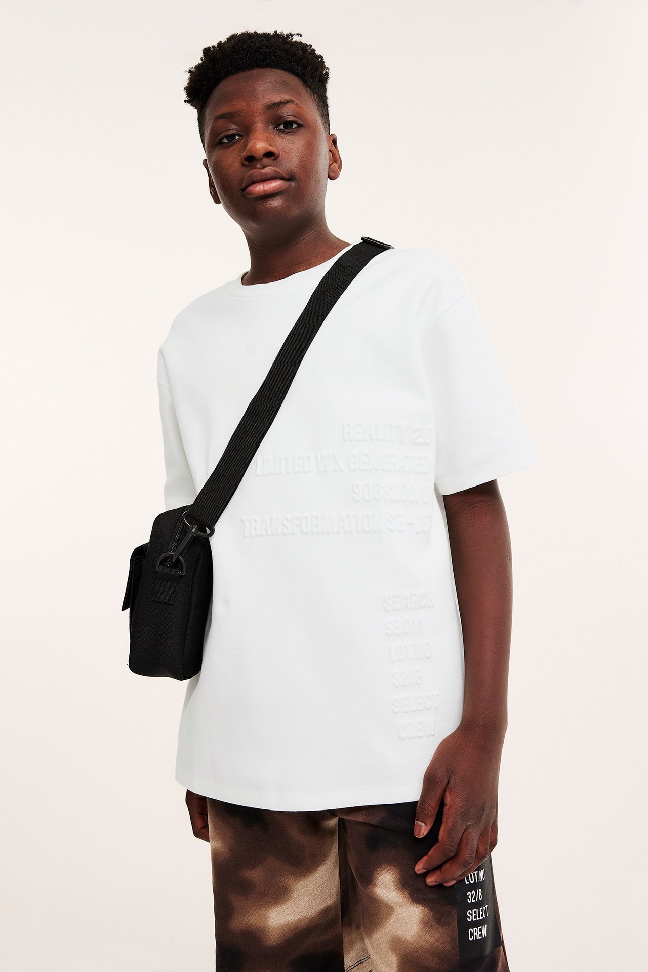 Widok z przodu: Dziecko w kremowym, oversize'owym T-shircie z wytłoczonym nadrukiem, brązowych szortach tie-dye i czarnej torbie.