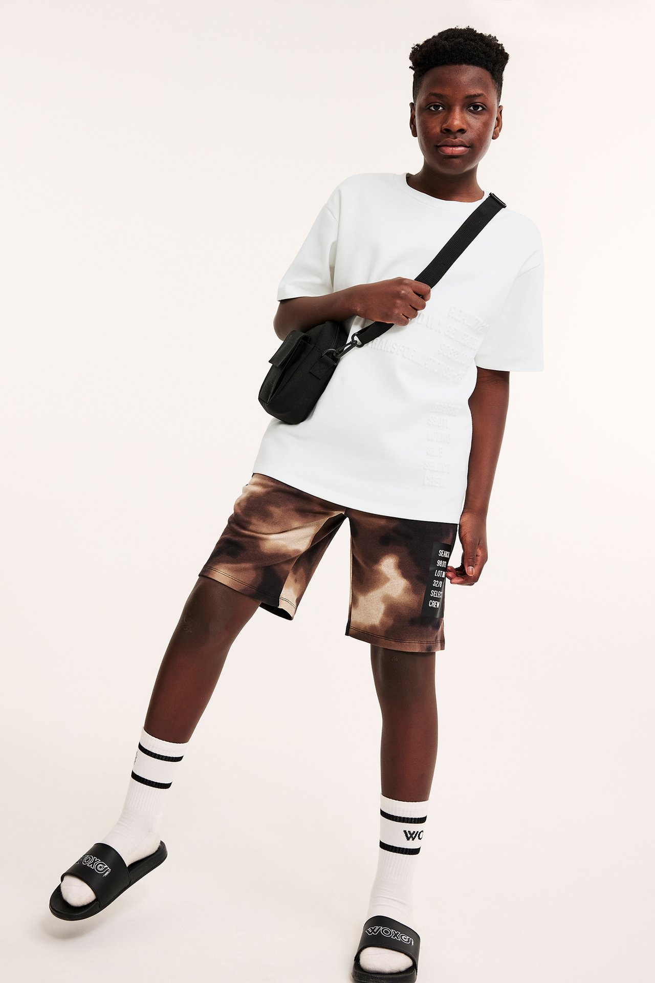 Framifrån: Barn i offwhite oversized t-shirt, bruna tie-dye shorts, vita strumpor, svarta tofflor, svart väska.