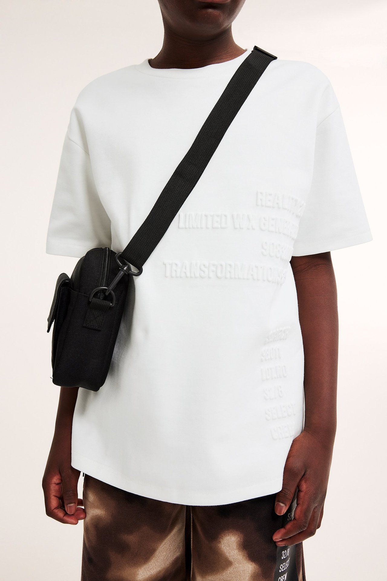 Widok z przodu: Dziecko w kremowym, oversize'owym bawełnianym T-shircie z wytłoczonym nadrukiem i czarną torbą.