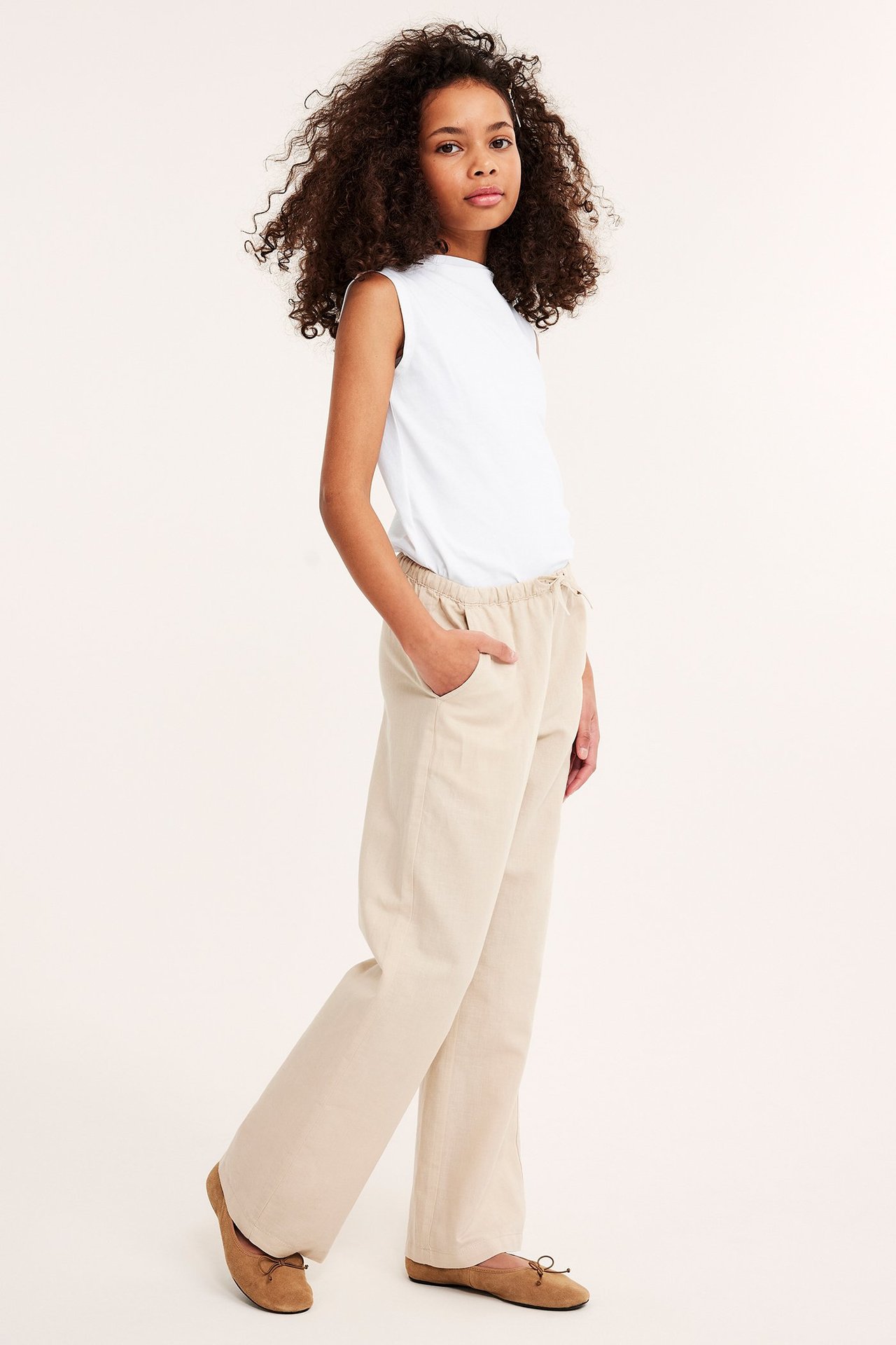 Sidovy: barn i vit ärmlös topp med båthals, beiga vida byxor och bruna ballerinaskor.