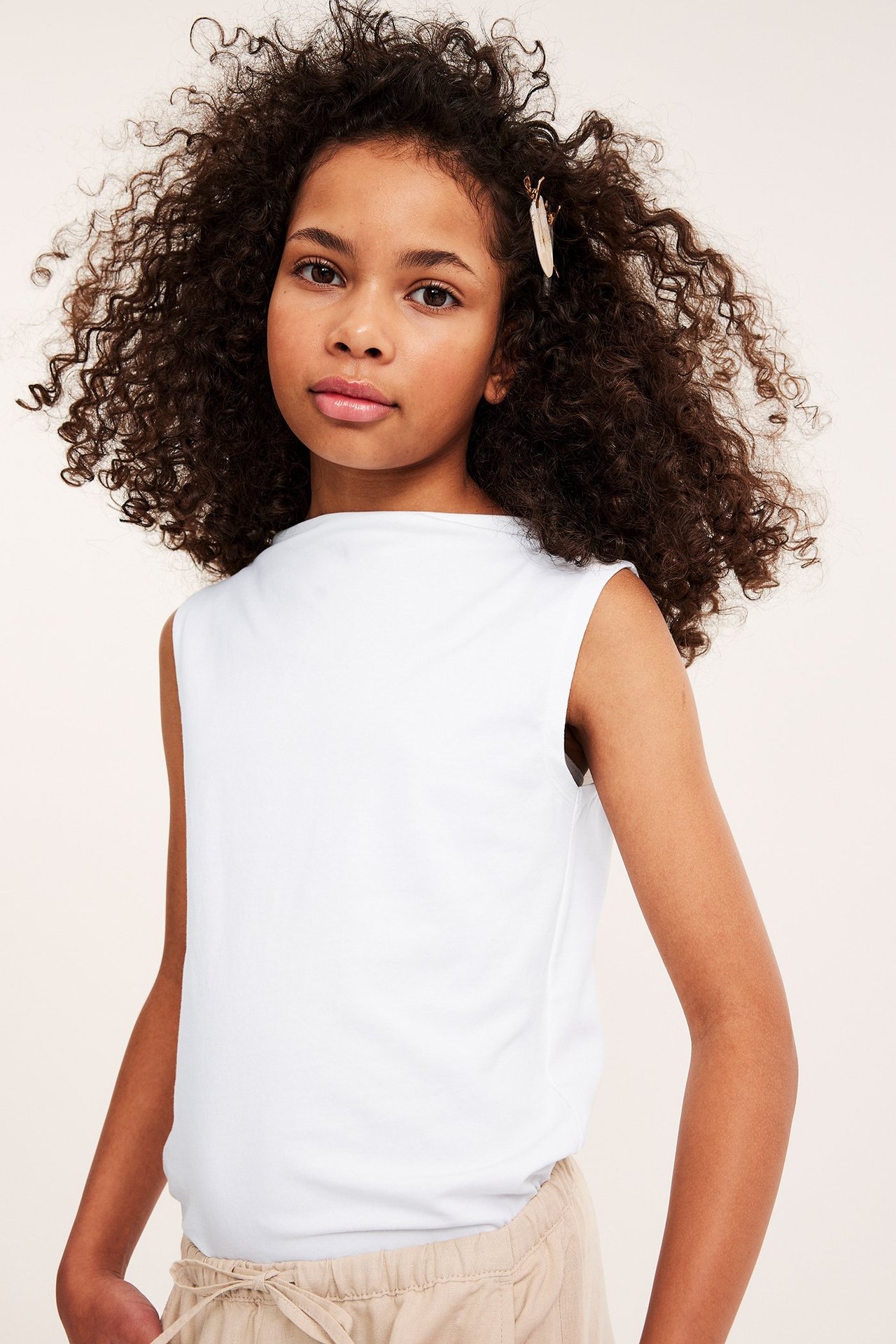 Framsida: Barn i vit ärmlös topp med båthals, beiga shorts och ett hårspänne.