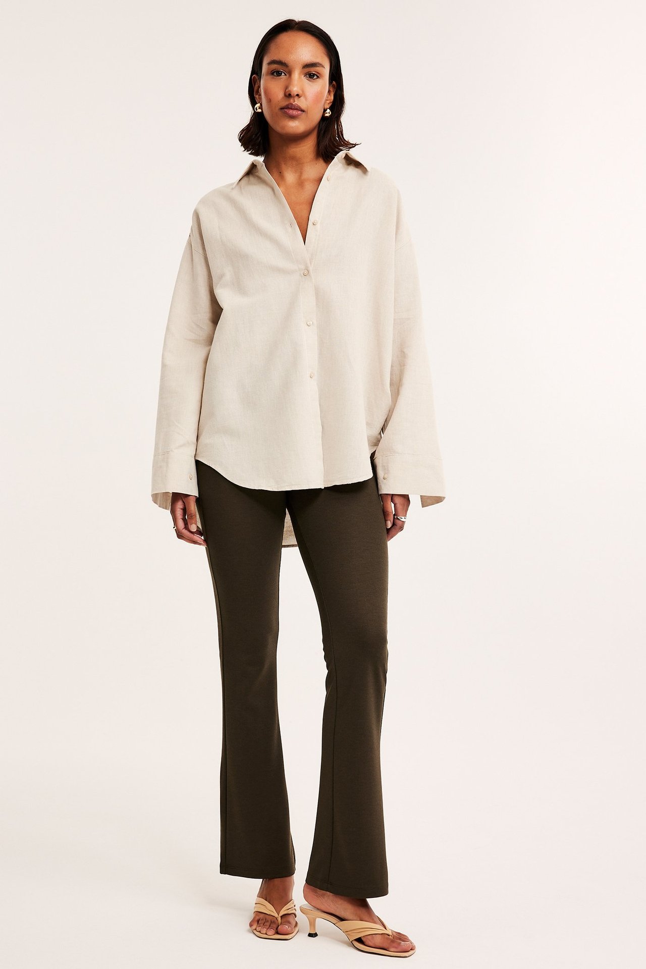 Widok z przodu: Kobieta w ciemnobrązowych, rozszerzanych spodniach dżersejowych, beżowej koszuli i sandałach.