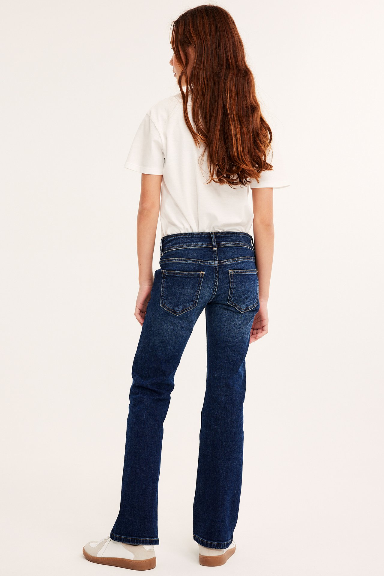Widok z tyłu: Dziecko w ciemnoniebieskich jeansach bootcut, białym T-shircie i beżowo-białych sneakersach.