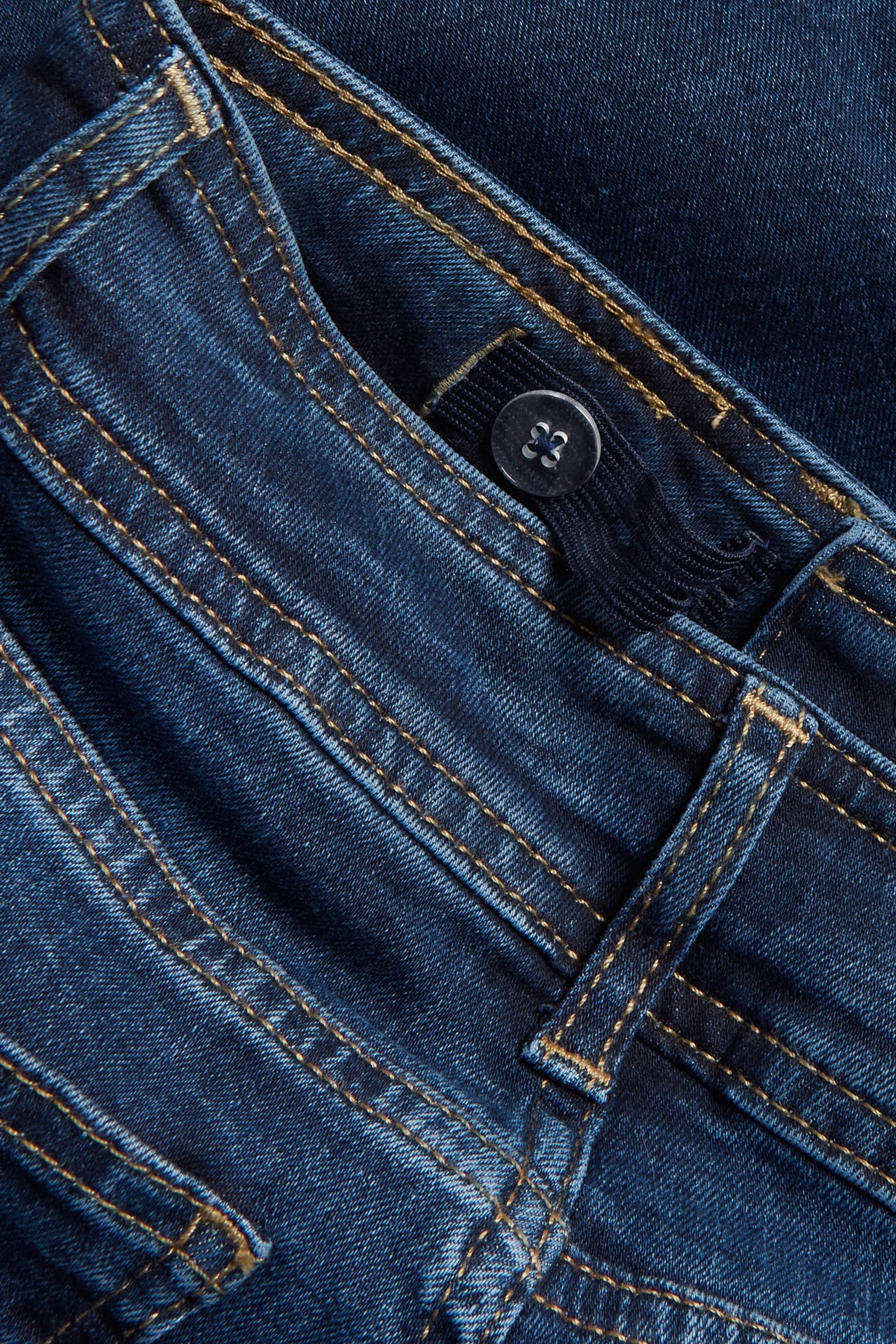 Zbliżenie na ciemnoniebieskie jeansy bootcut z pomarańczowymi przeszyciami i regulowanym guzikiem w pasie.