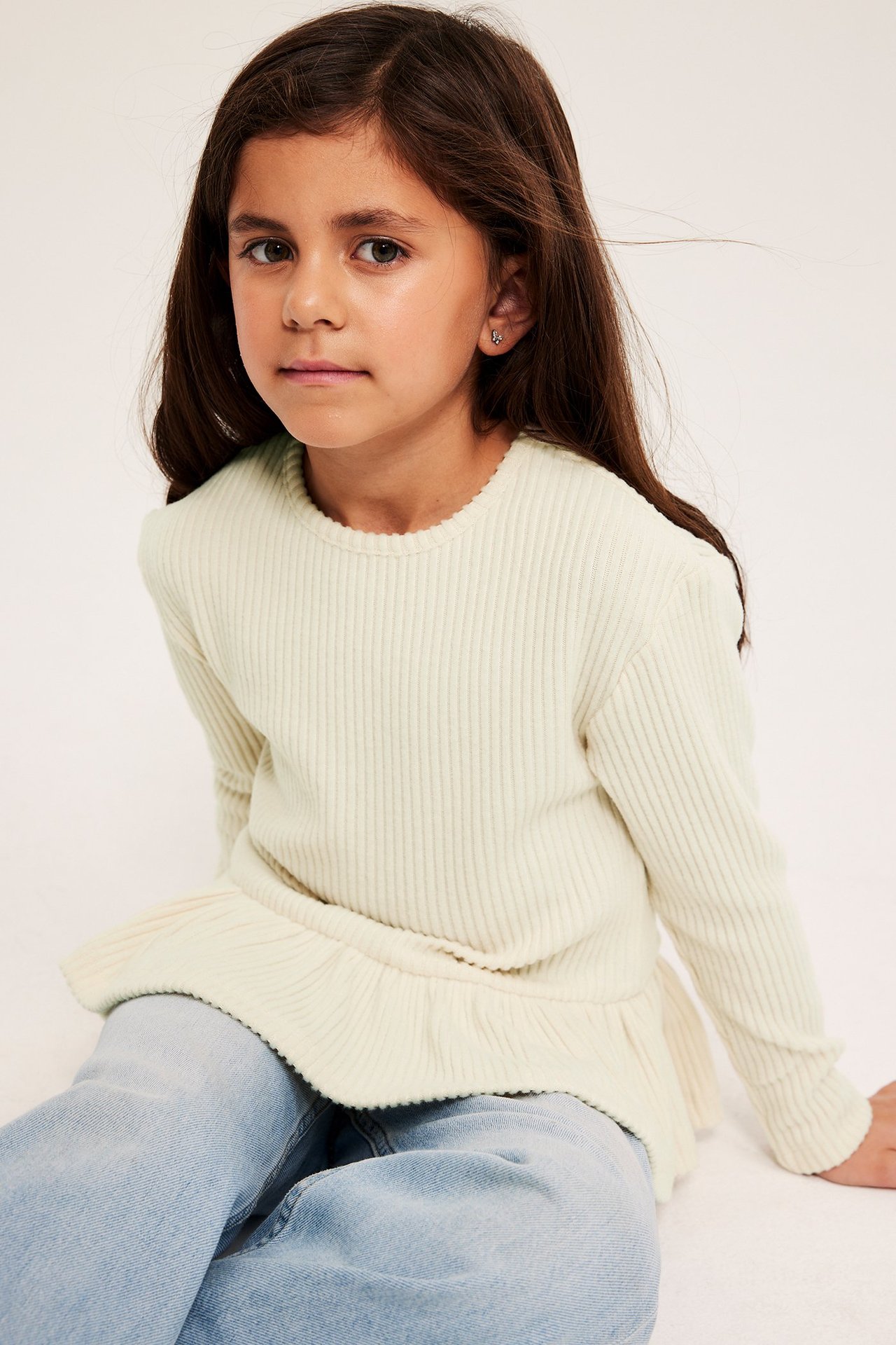 Frontbilde av et barn i en lys beige ribbet langermet peplumtopp over lyseblå jeans.